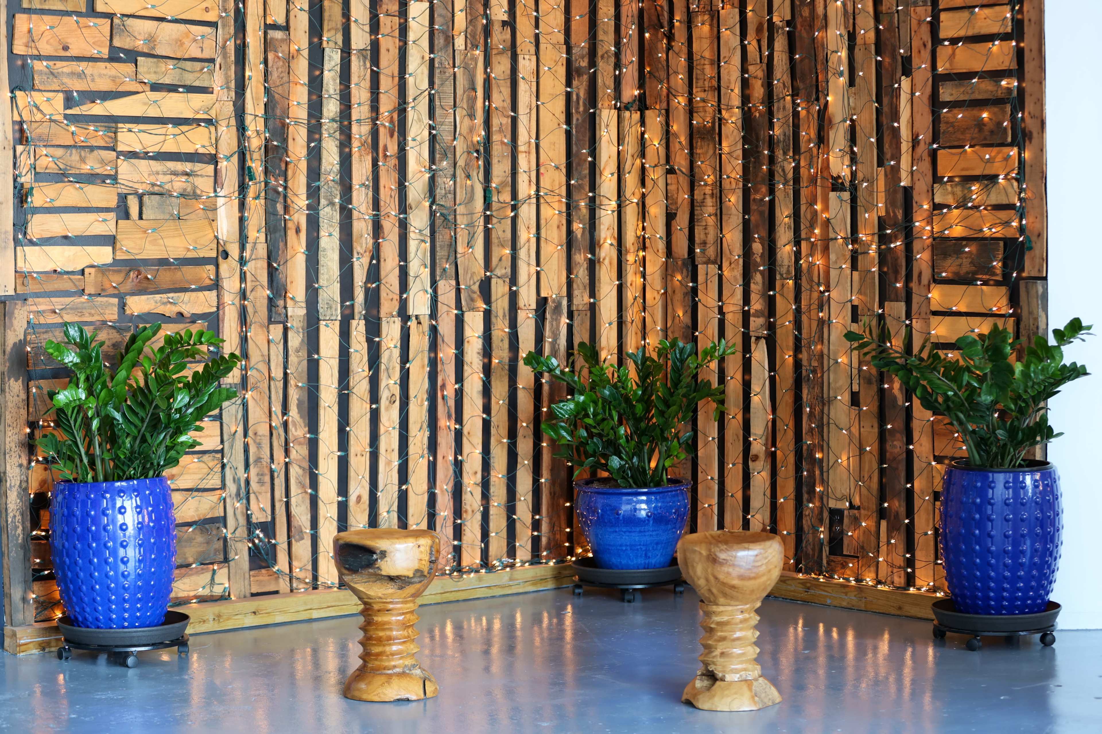 The image shows a decorated interior space with a wooden feature wall, three potted plants in blue containers, and two wooden stools arranged on a polished floor.