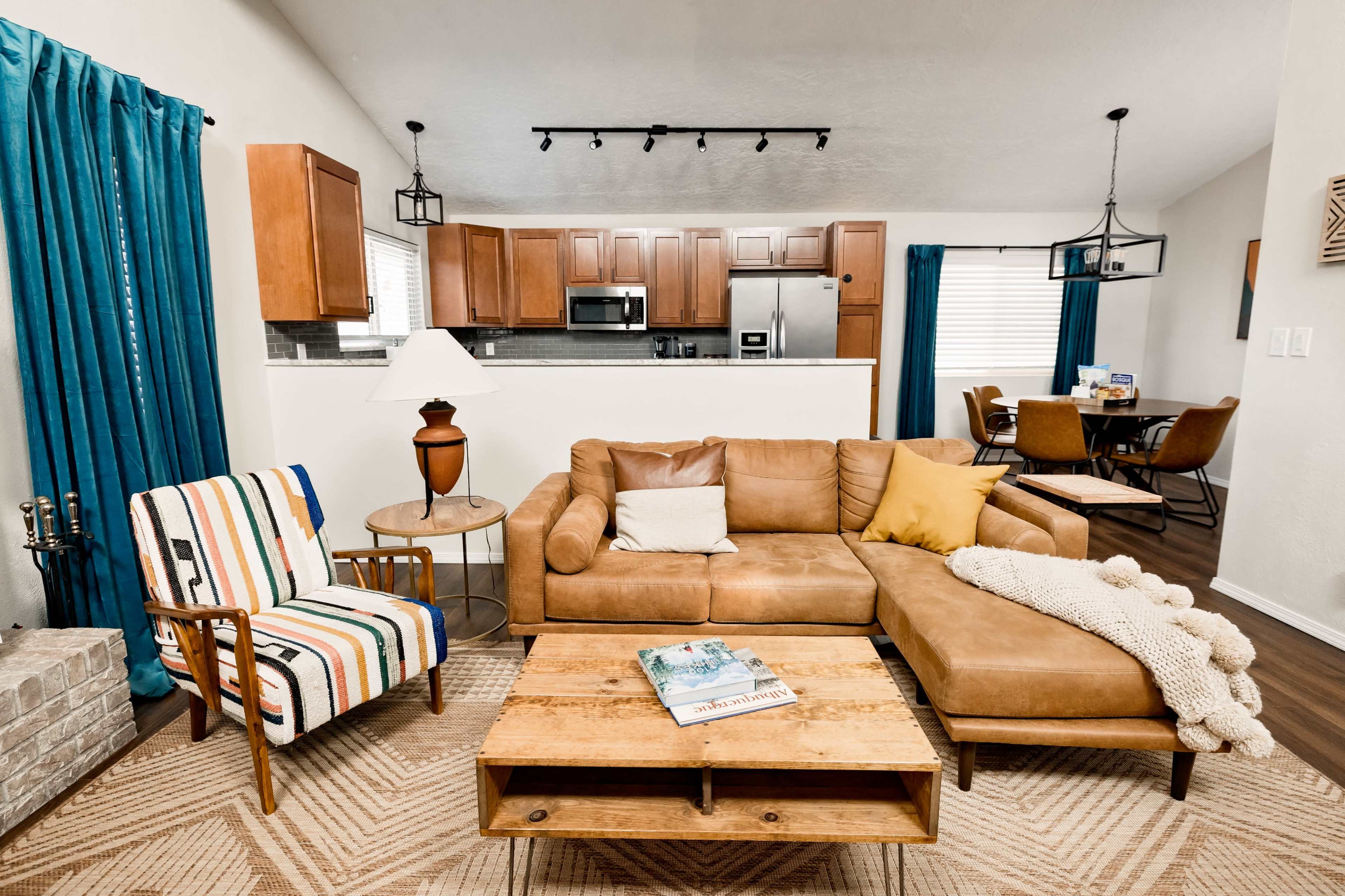 A cozy living room features a brown leather couch, a striped armchair, a coffee table with a magazine, and a kitchen area visible in the background.