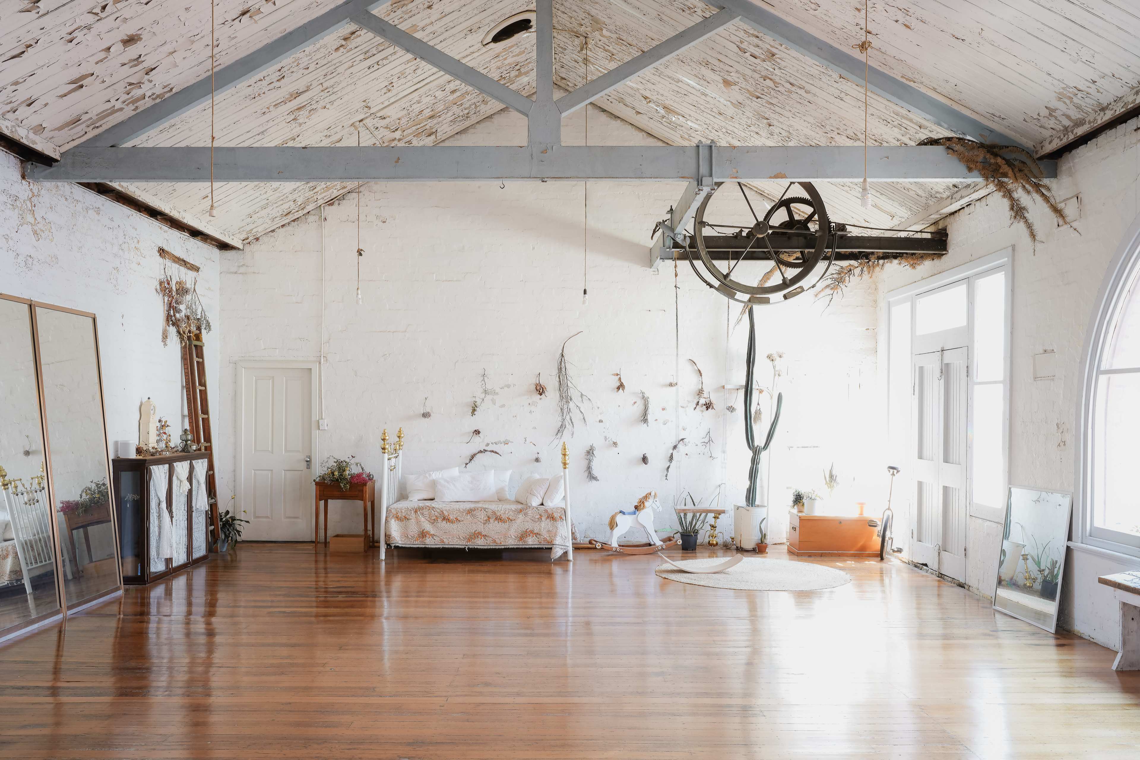 The image showcases a spacious, rustic interior with exposed beams, wooden flooring, and minimal furnishings, including a bed and a large mirror.