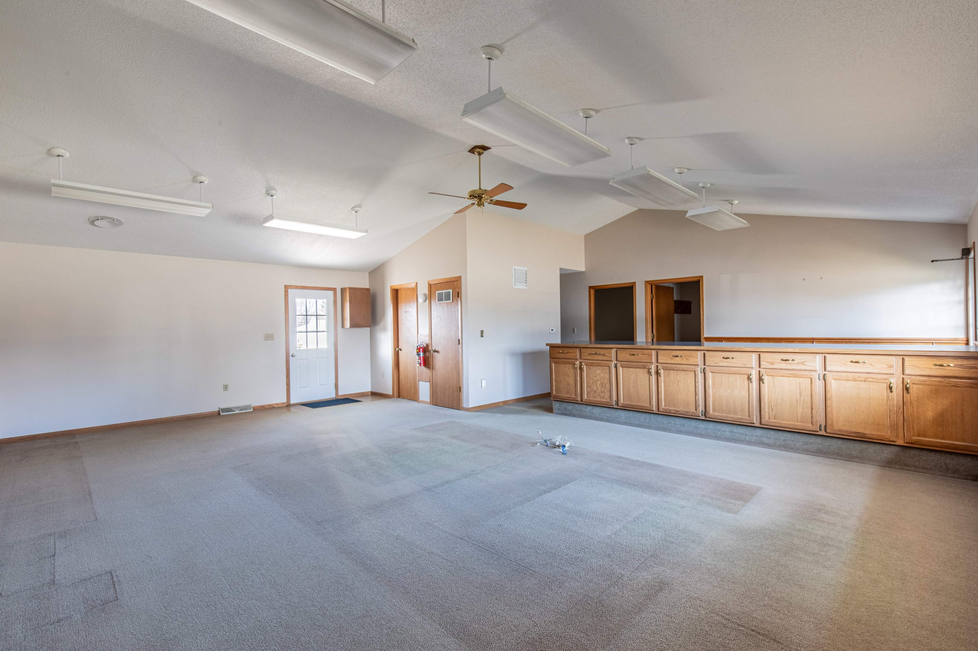 A spacious, empty room with a ceiling fan, overhead lights, and a row of wooden cabinets along one wall.