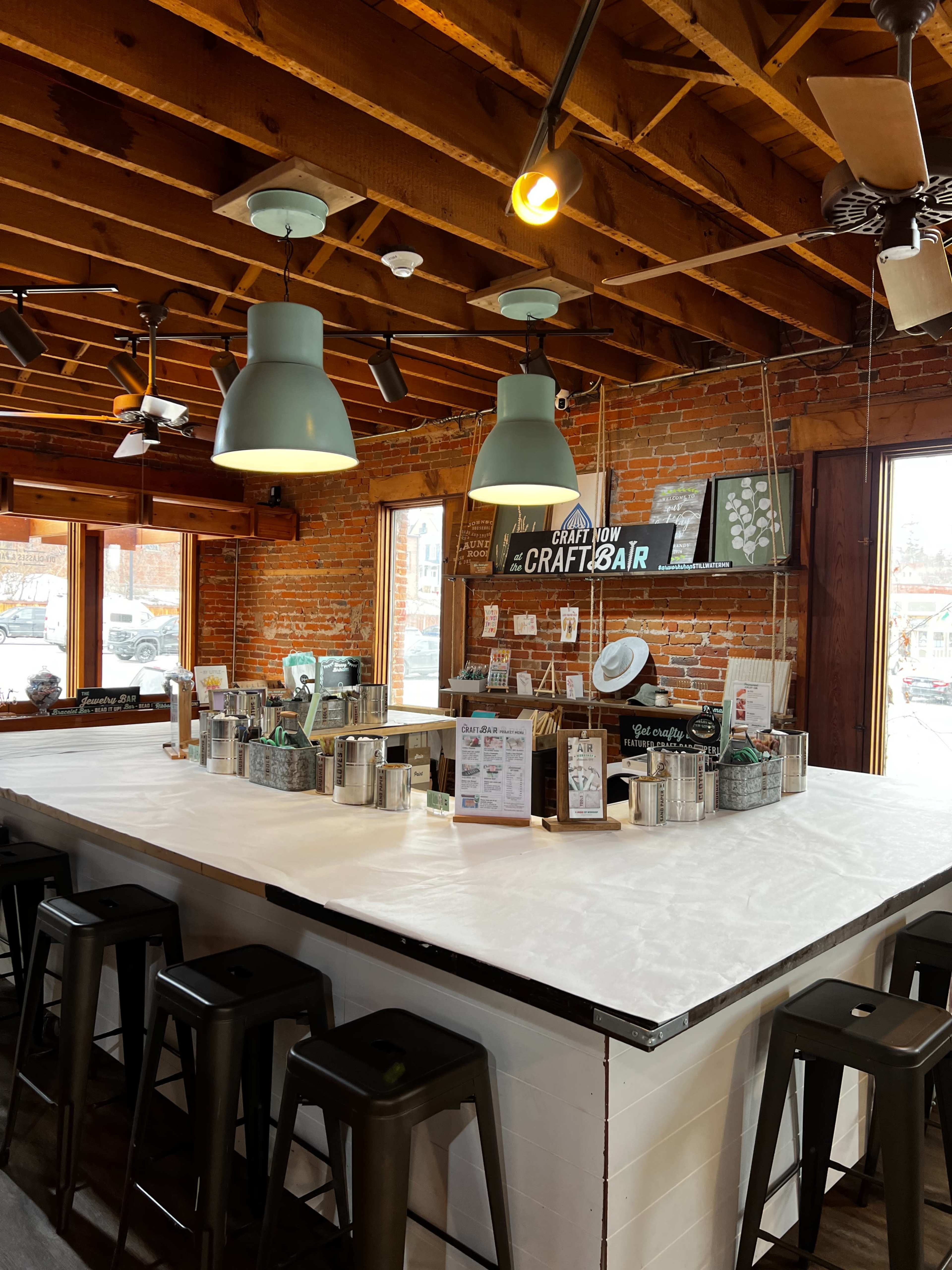The image shows a well-lit bar area with a white countertop, metal stools, exposed brick walls, and pendant lighting hanging from a wooden ceiling.