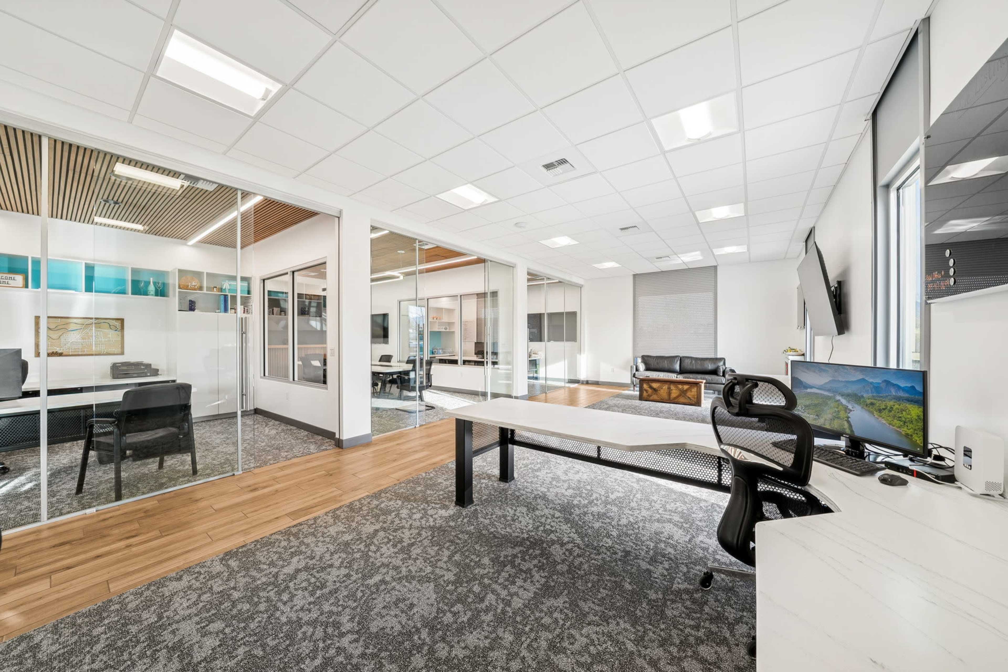 The image shows a modern office space featuring a large desk, a black ergonomic chair, and glass-walled conference rooms in the background.