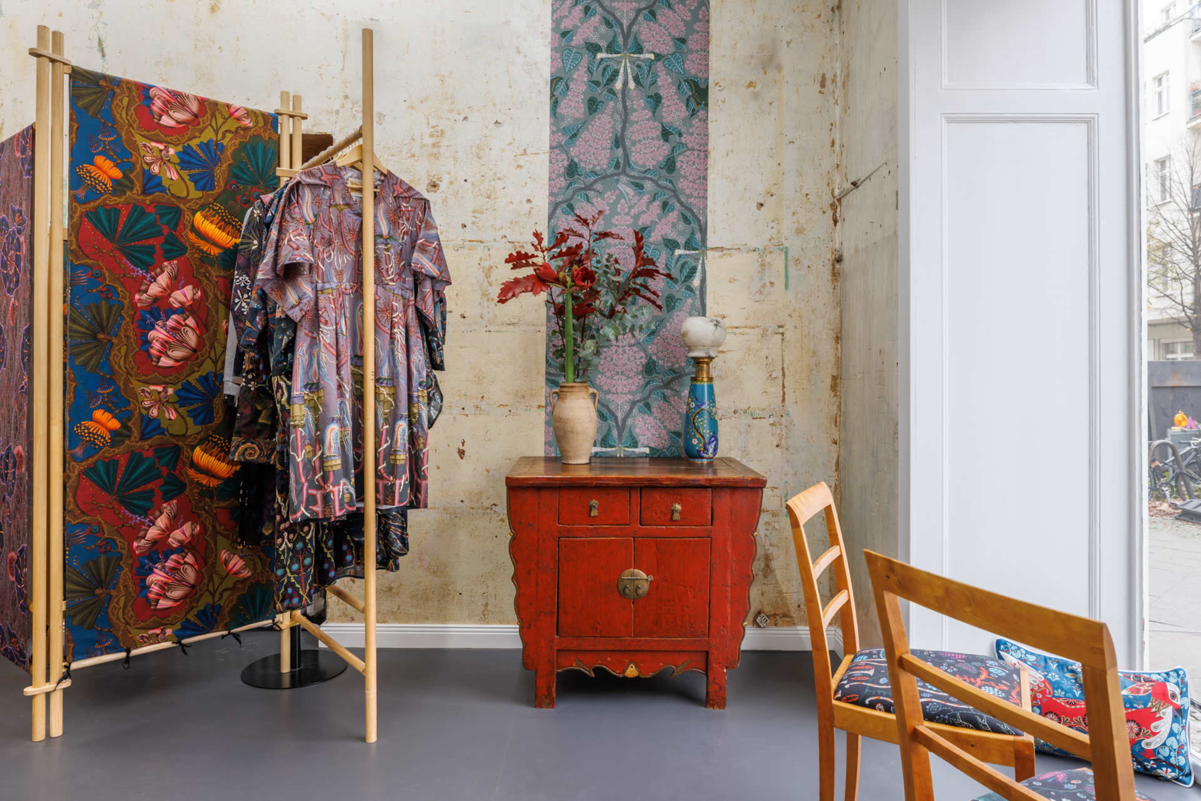 A retail space featuring colorful clothing displayed on a rack, a red wooden cabinet, and a decorative vase with flowers against a textured wall.