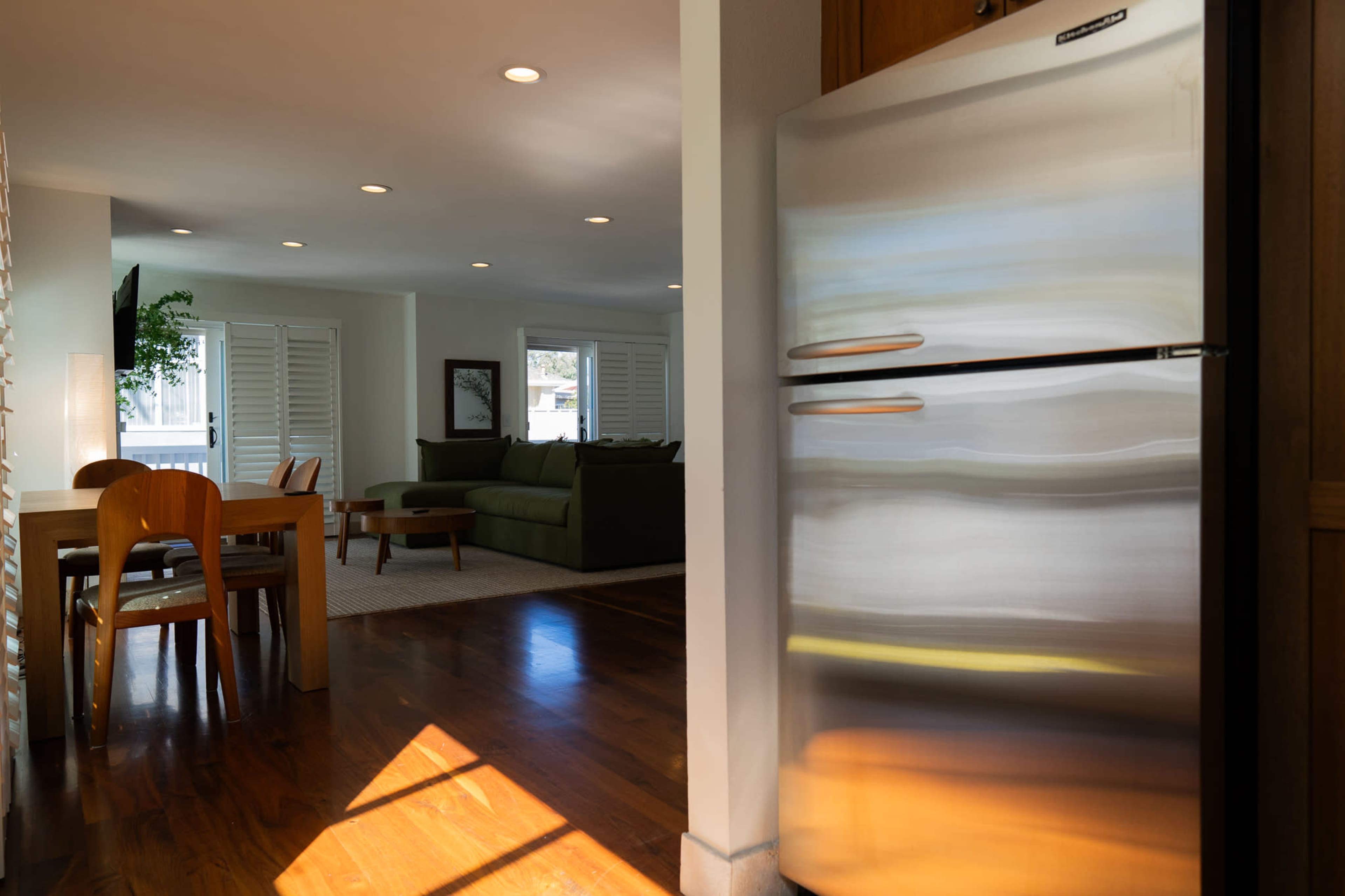 The image shows a modern kitchen and living area with a stainless steel refrigerator, wooden dining table, and a green sofa facing a television.