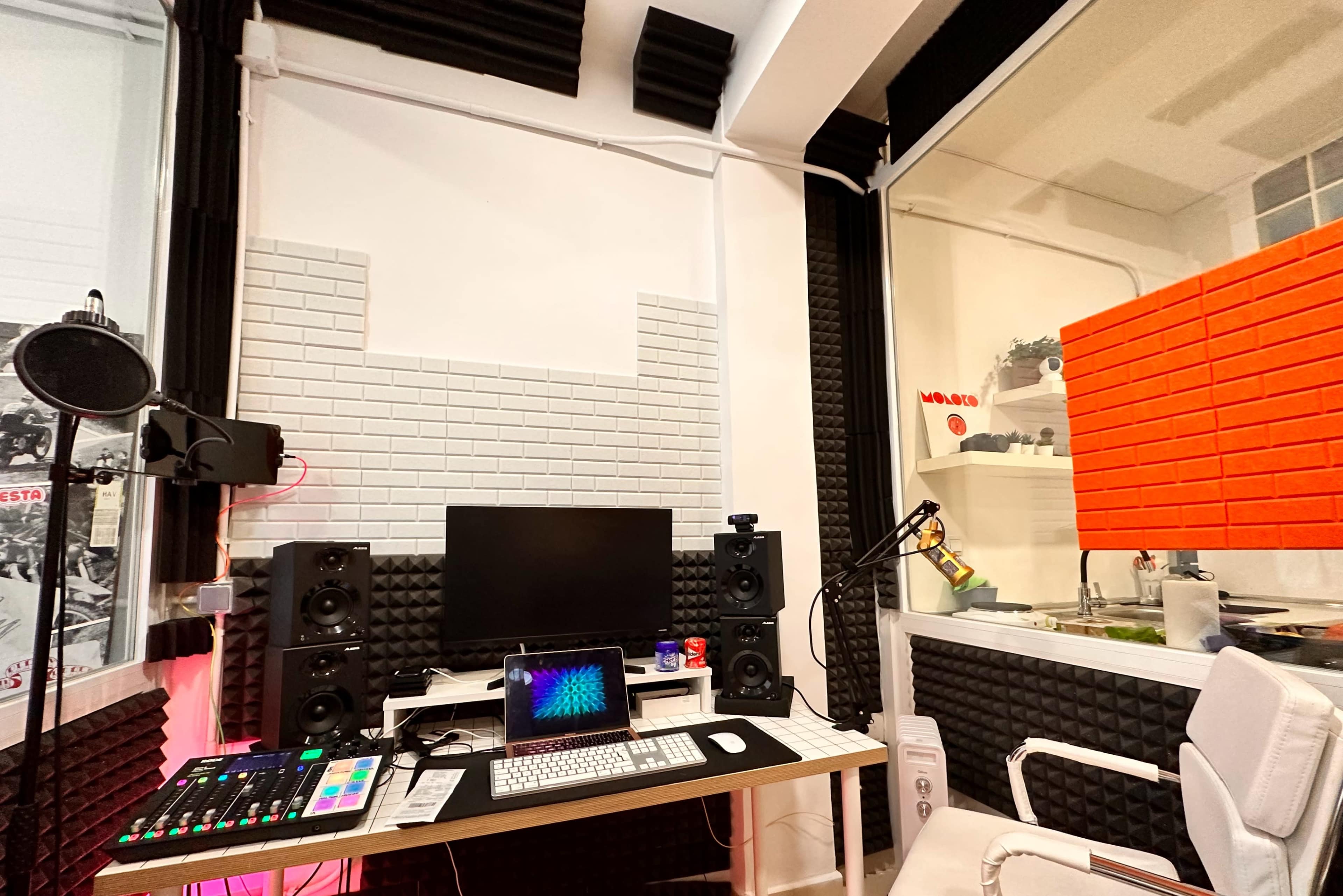 A recording studio setup featuring a microphone, computer monitor, mixing console, and a desk with colorful soundproofing panels.