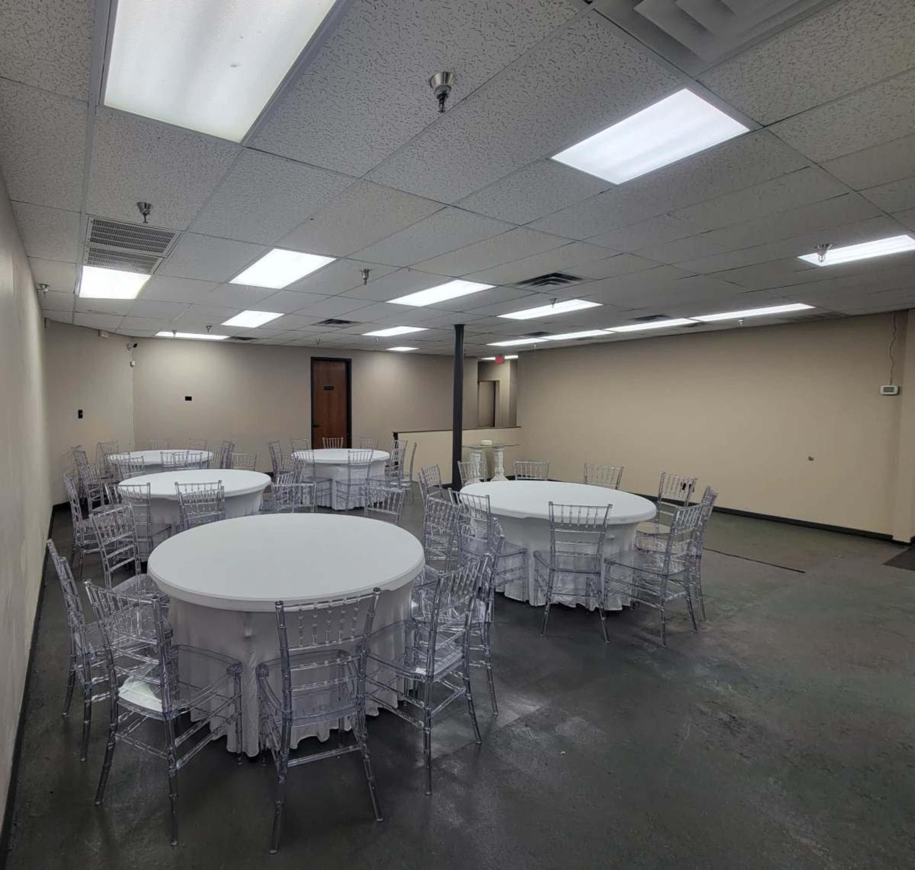 The image shows a spacious indoor venue with several round tables covered by white tablecloths and surrounded by transparent chairs.