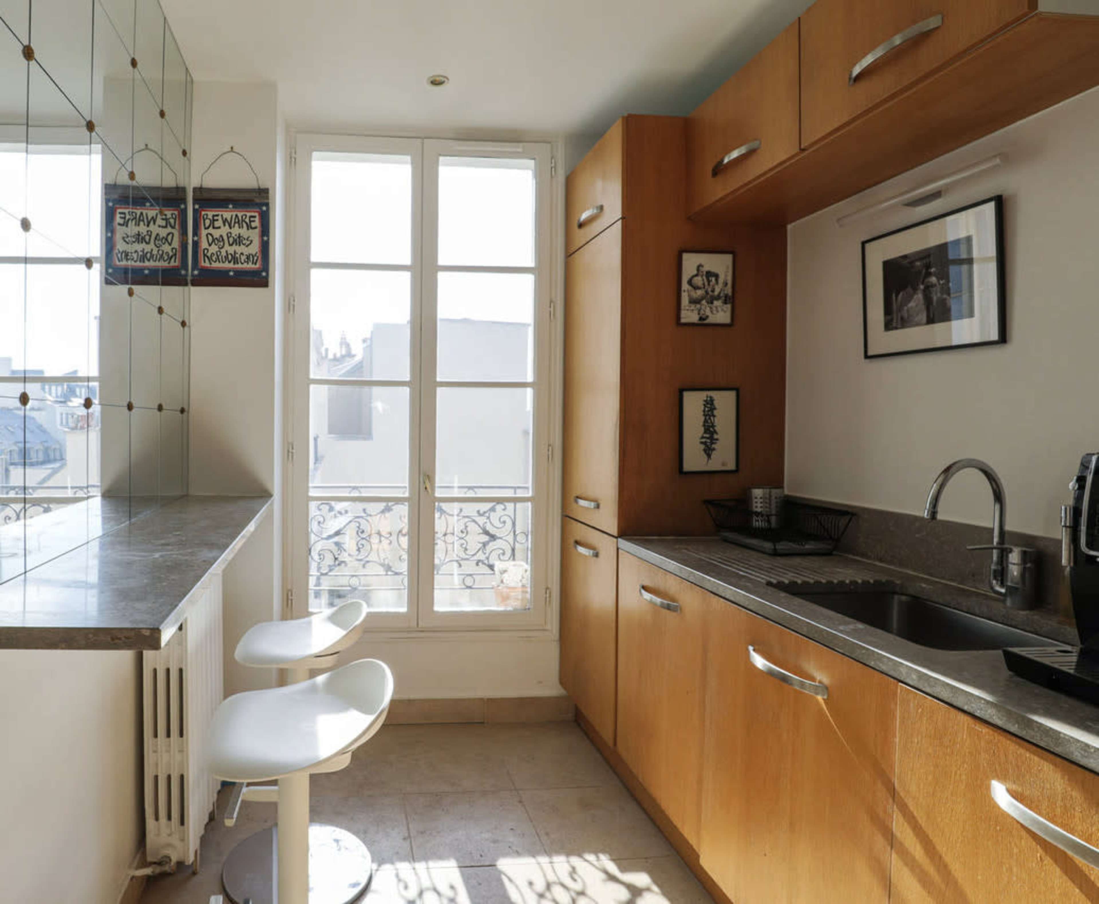 A modern kitchen features wooden cabinetry, a stainless steel sink, and two white stools at a counter near a window with a view.