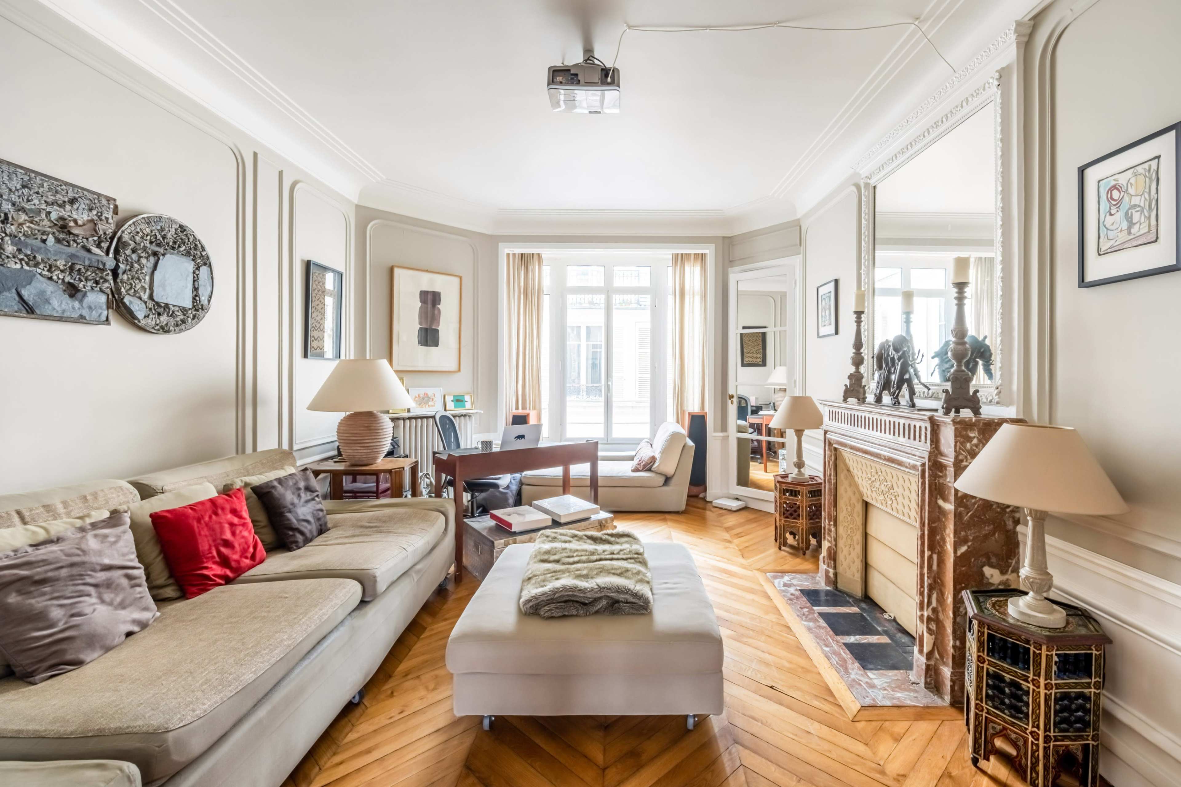 The image shows a well-furnished living room with a beige sofa, a coffee table, and a decorative fireplace surrounded by artwork and large windows letting in natural light.
