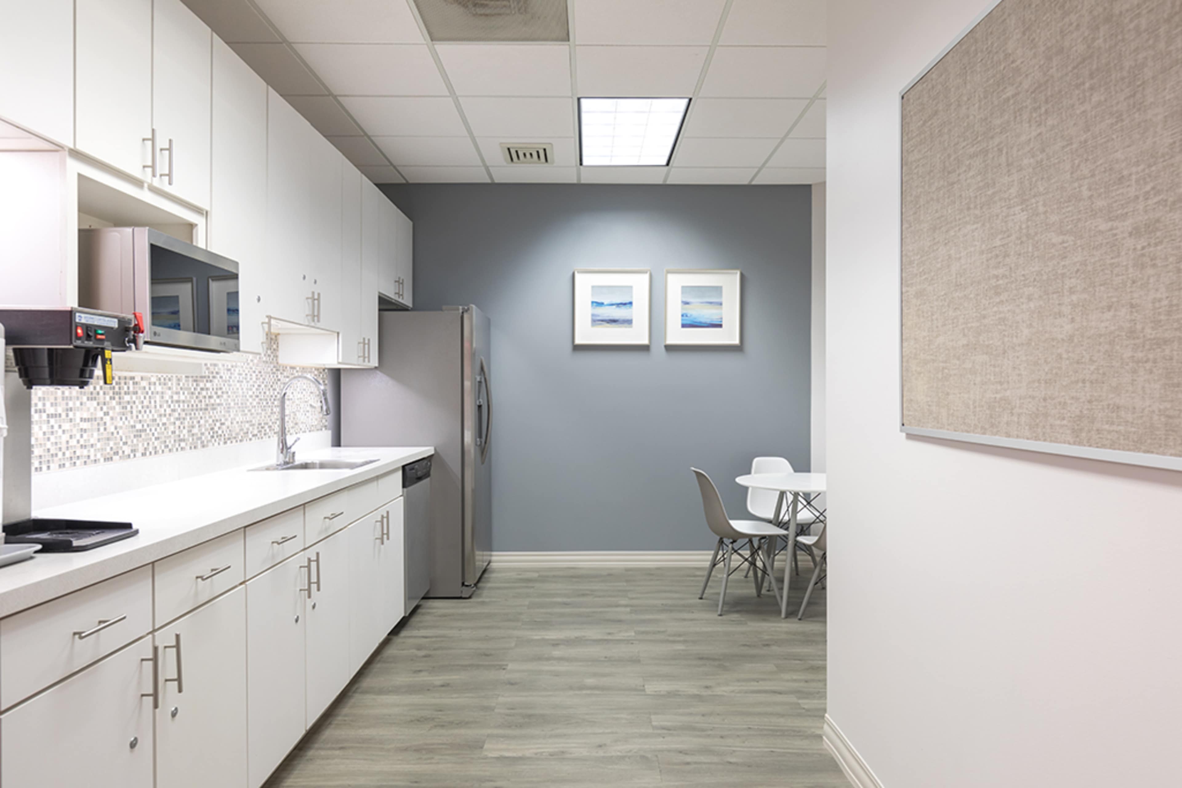 The image depicts a modern kitchen area with white cabinetry, stainless steel appliances, a small dining table, and light grey walls adorned with two framed pictures.