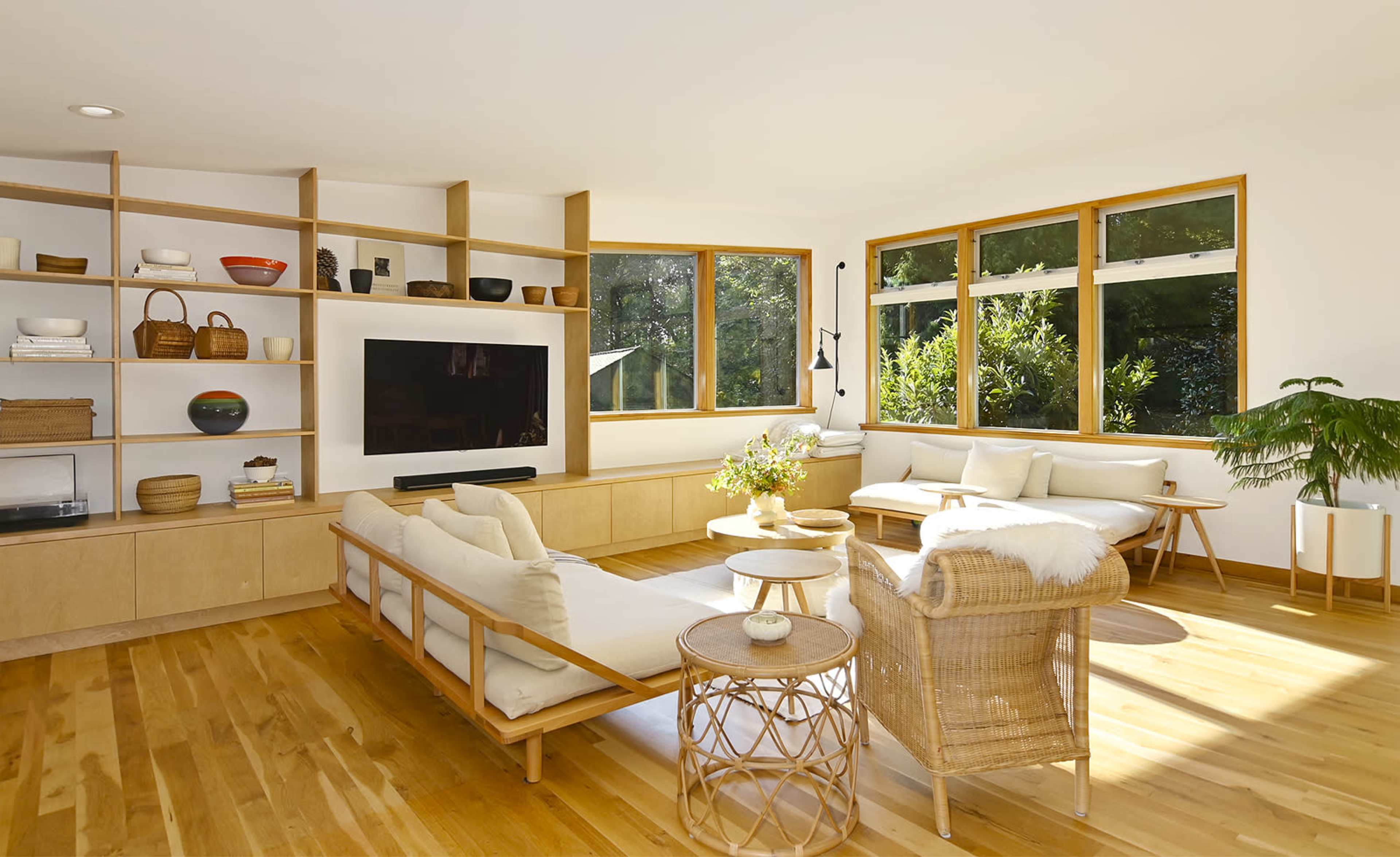 The room features a light-filled living area with wooden flooring, a minimalist shelf display, a television, and comfortable seating arrangements.