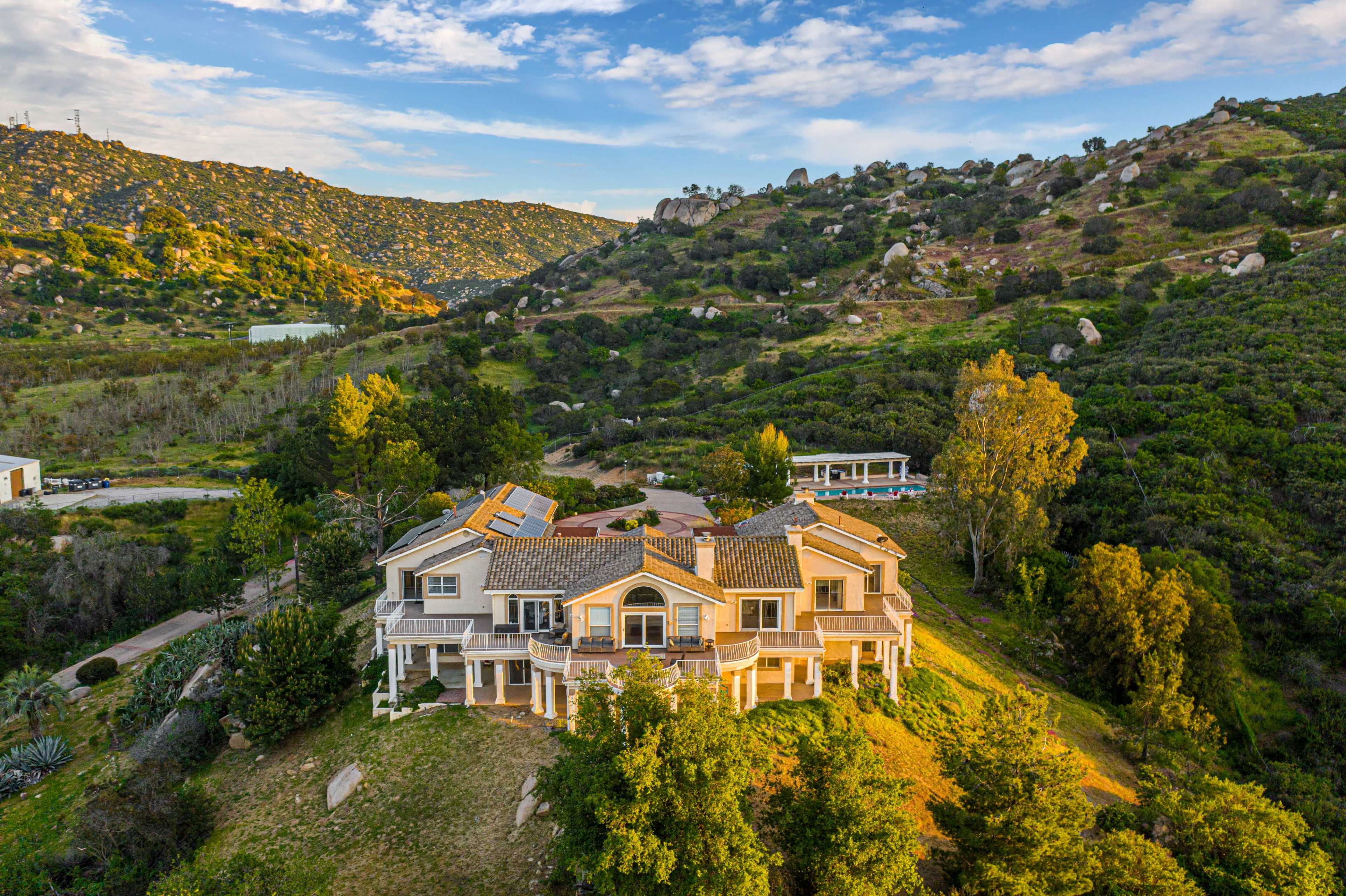 A large, multi-story house is situated on a hillside surrounded by greenery and rocky terrain under a partly cloudy sky.