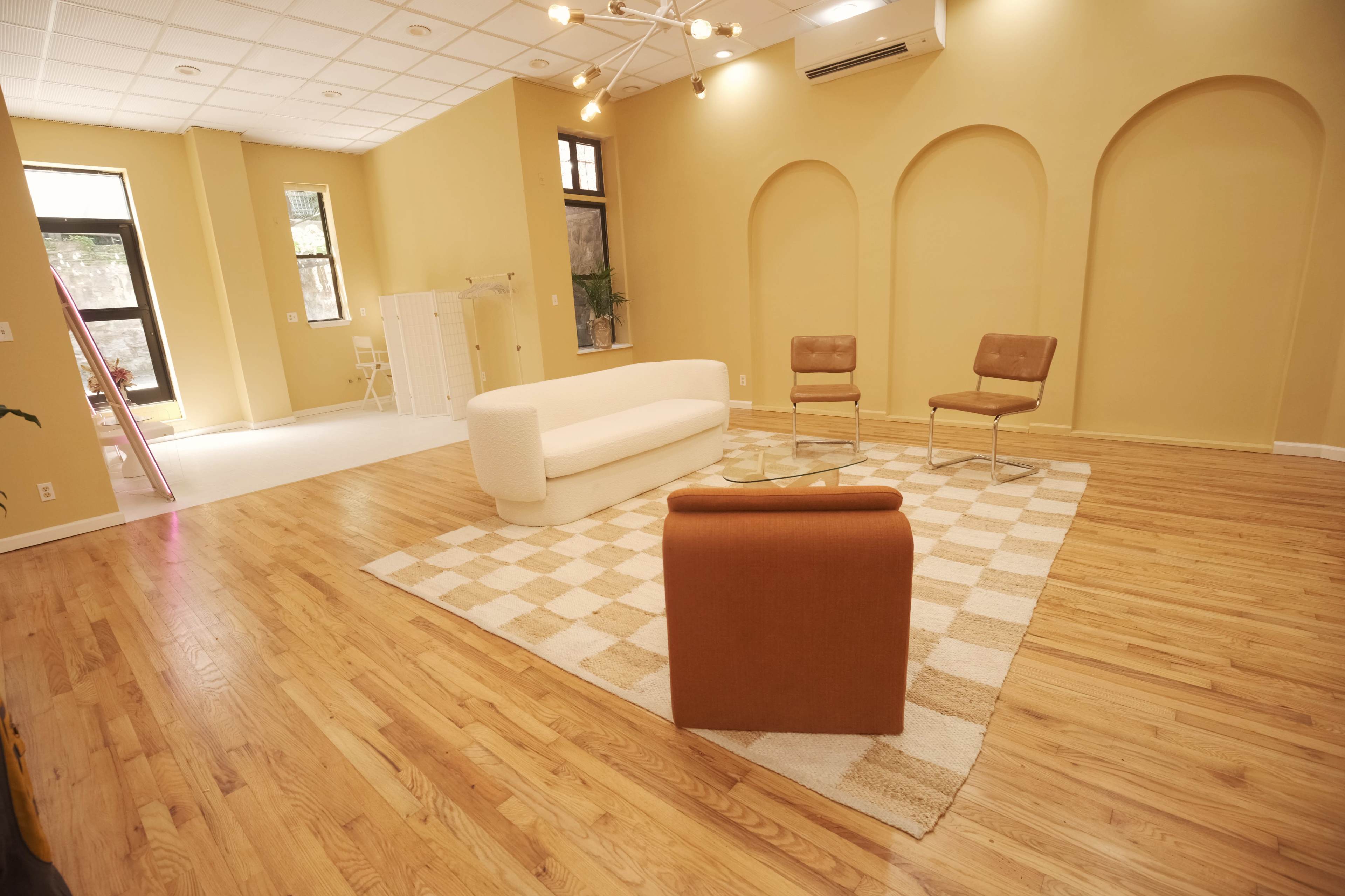 The image shows a spacious, light-filled room with wooden flooring, featuring a white sofa, a brown armchair, two brown chairs, and a patterned rug.