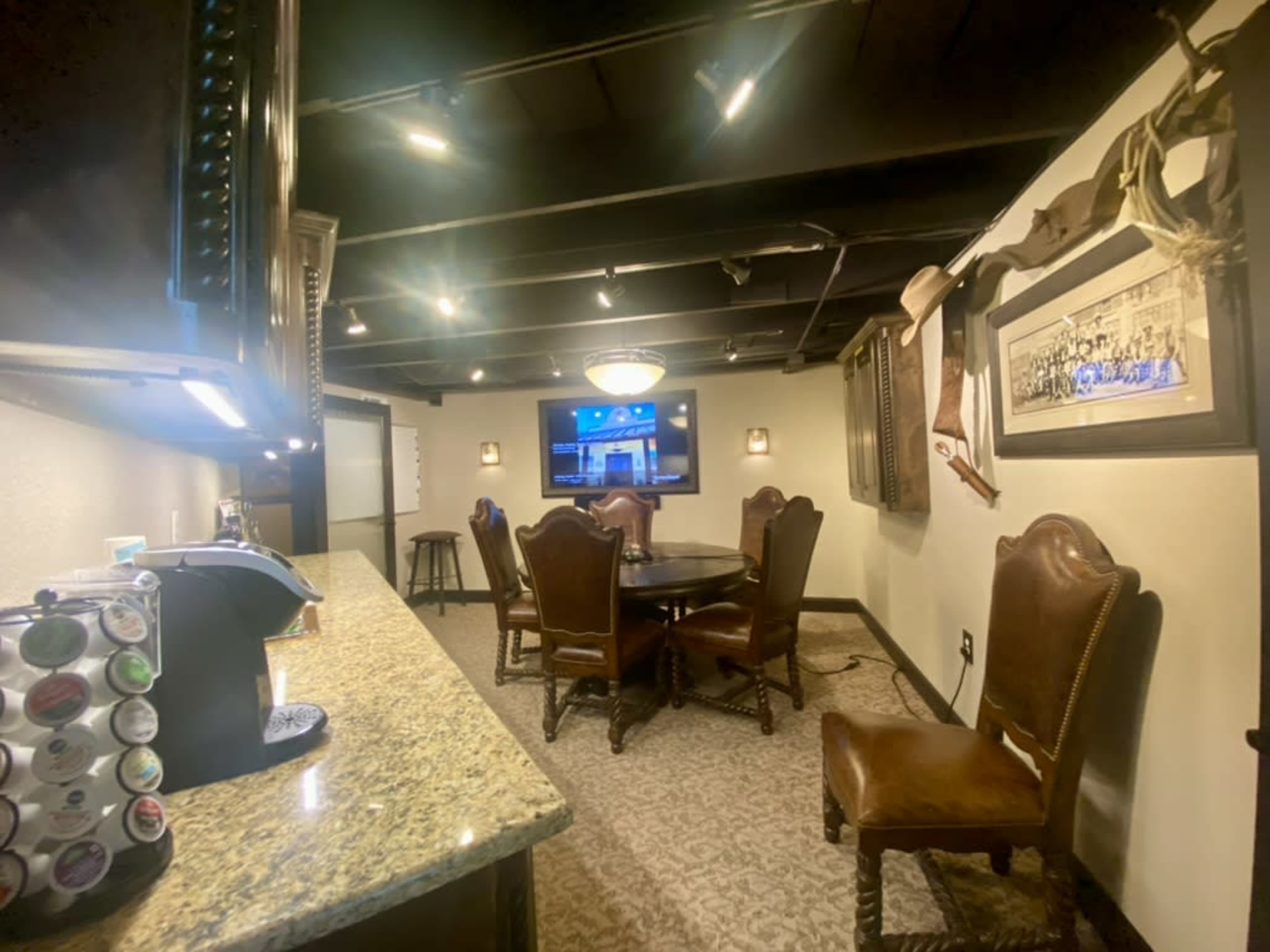 The image shows a dimly lit room featuring a small bar area with a granite countertop, a round wooden dining table surrounded by chairs, and a television mounted on the wall.