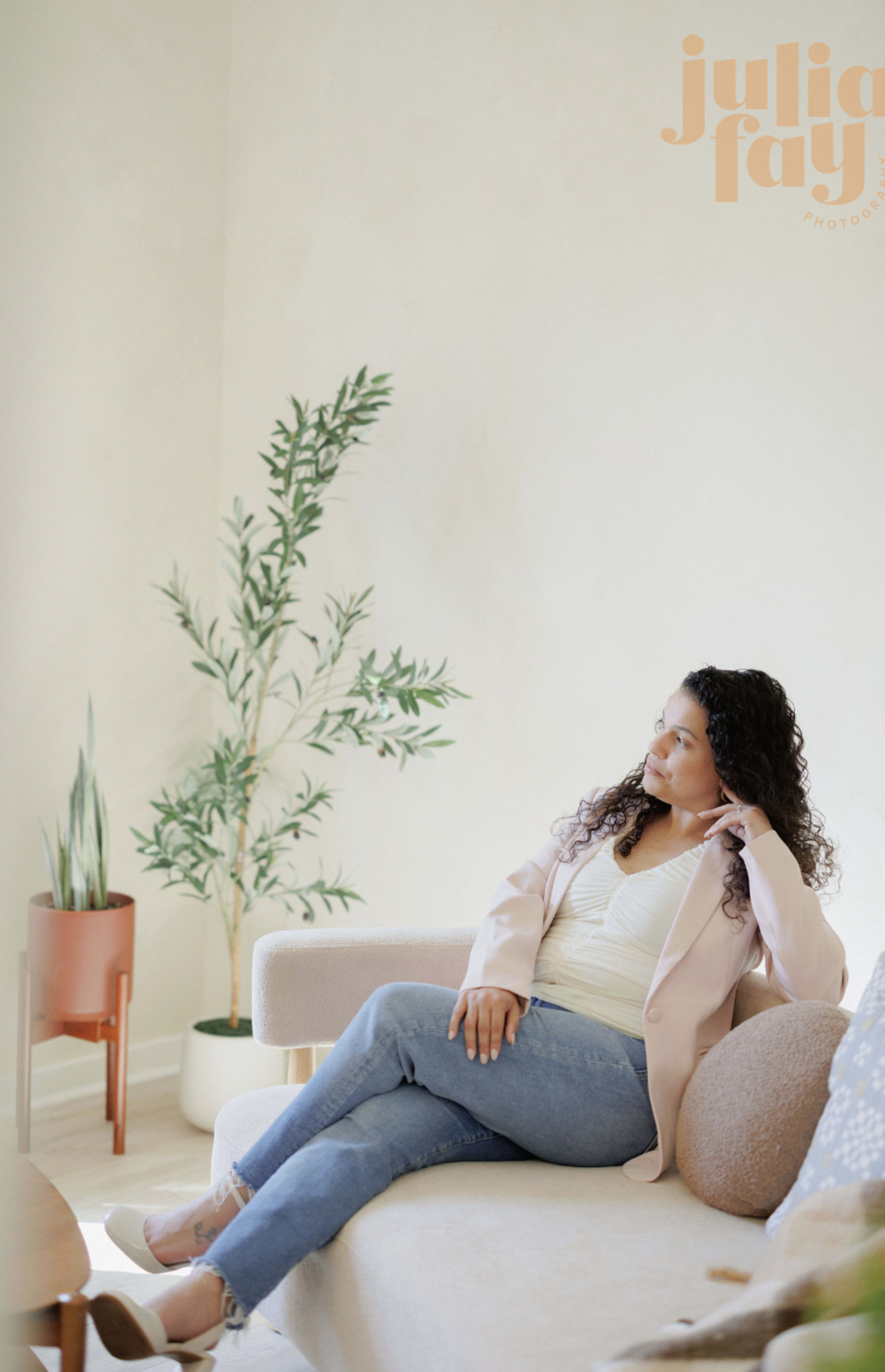 A woman with curly hair sits on a beige sofa in a softly lit room adorned with plants.