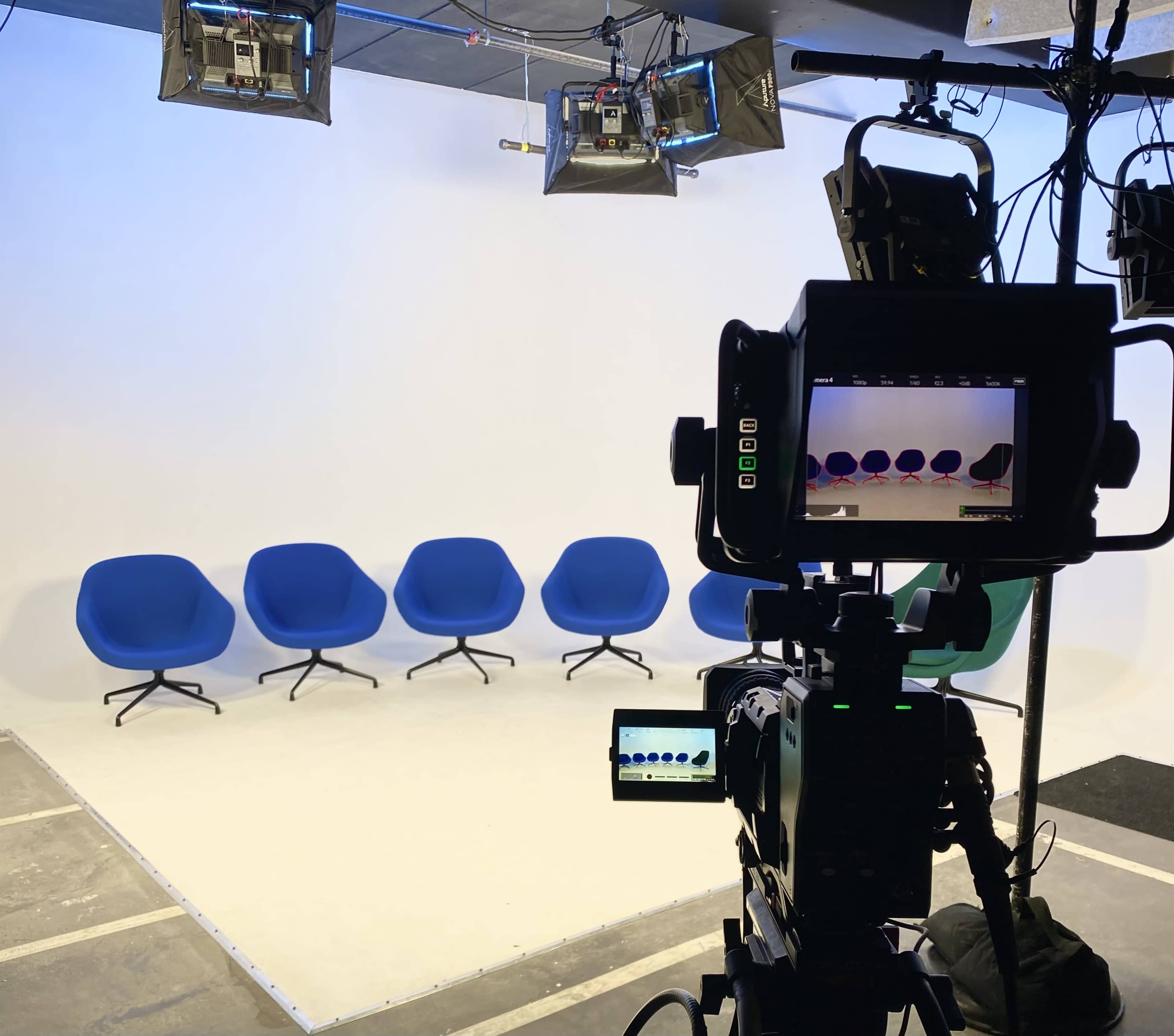 A camera setup faces a row of five blue chairs against a white backdrop in a studio.