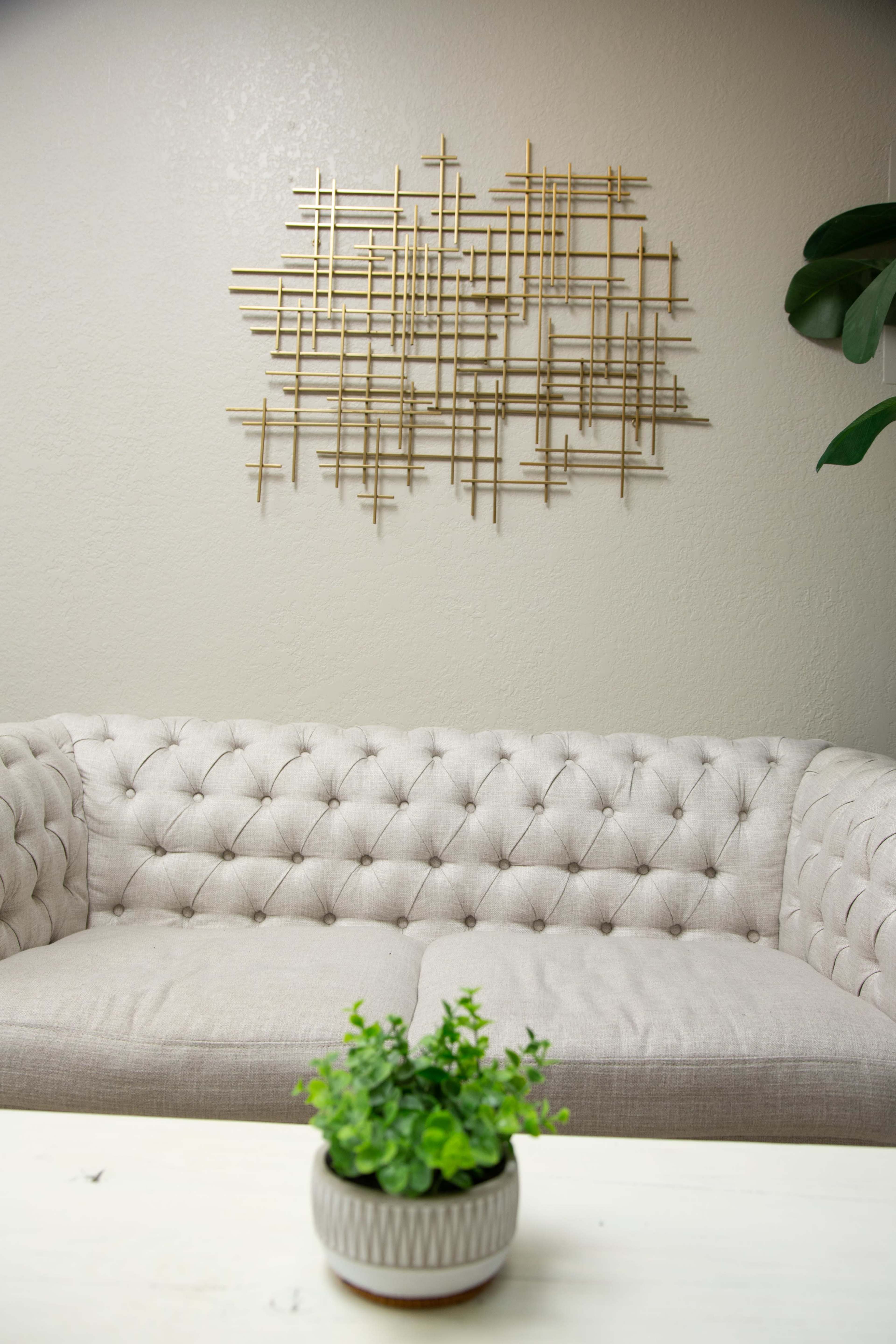 A light-colored tufted sofa sits below a gold geometric wall art piece, with a small potted plant on a white table in front.