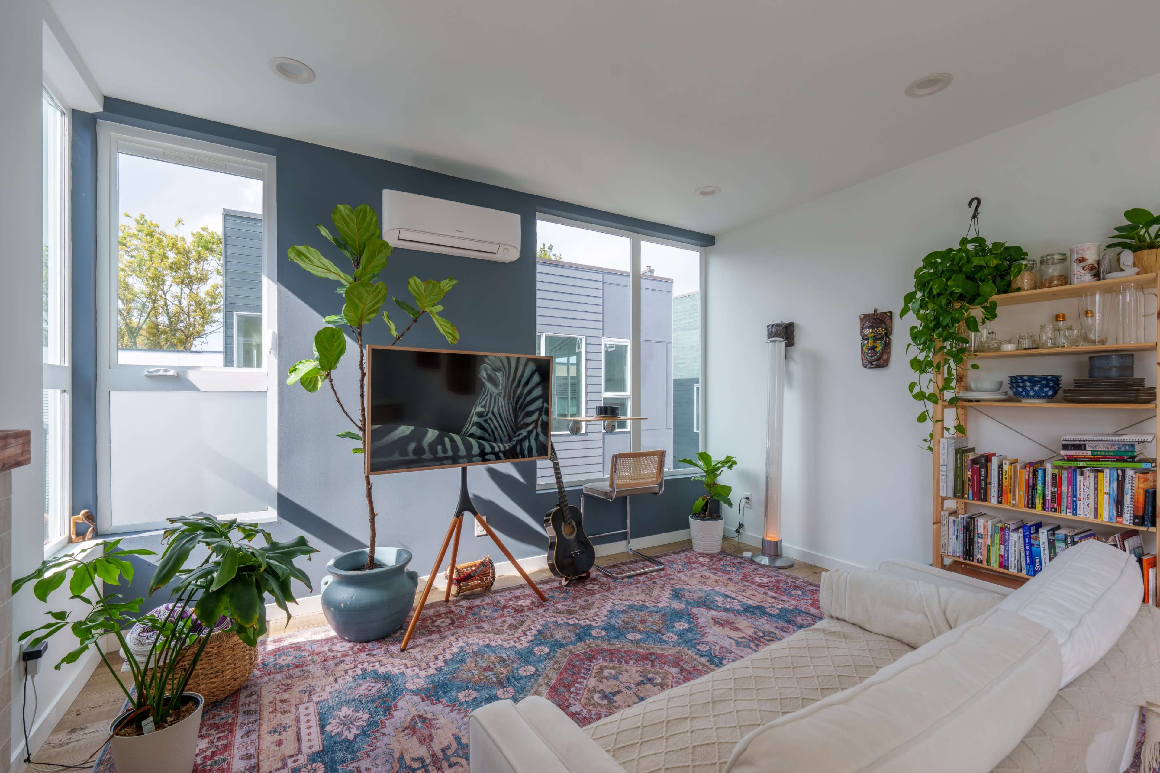A bright living room featuring a large television, a white couch, a bookshelf filled with books, and several indoor plants.