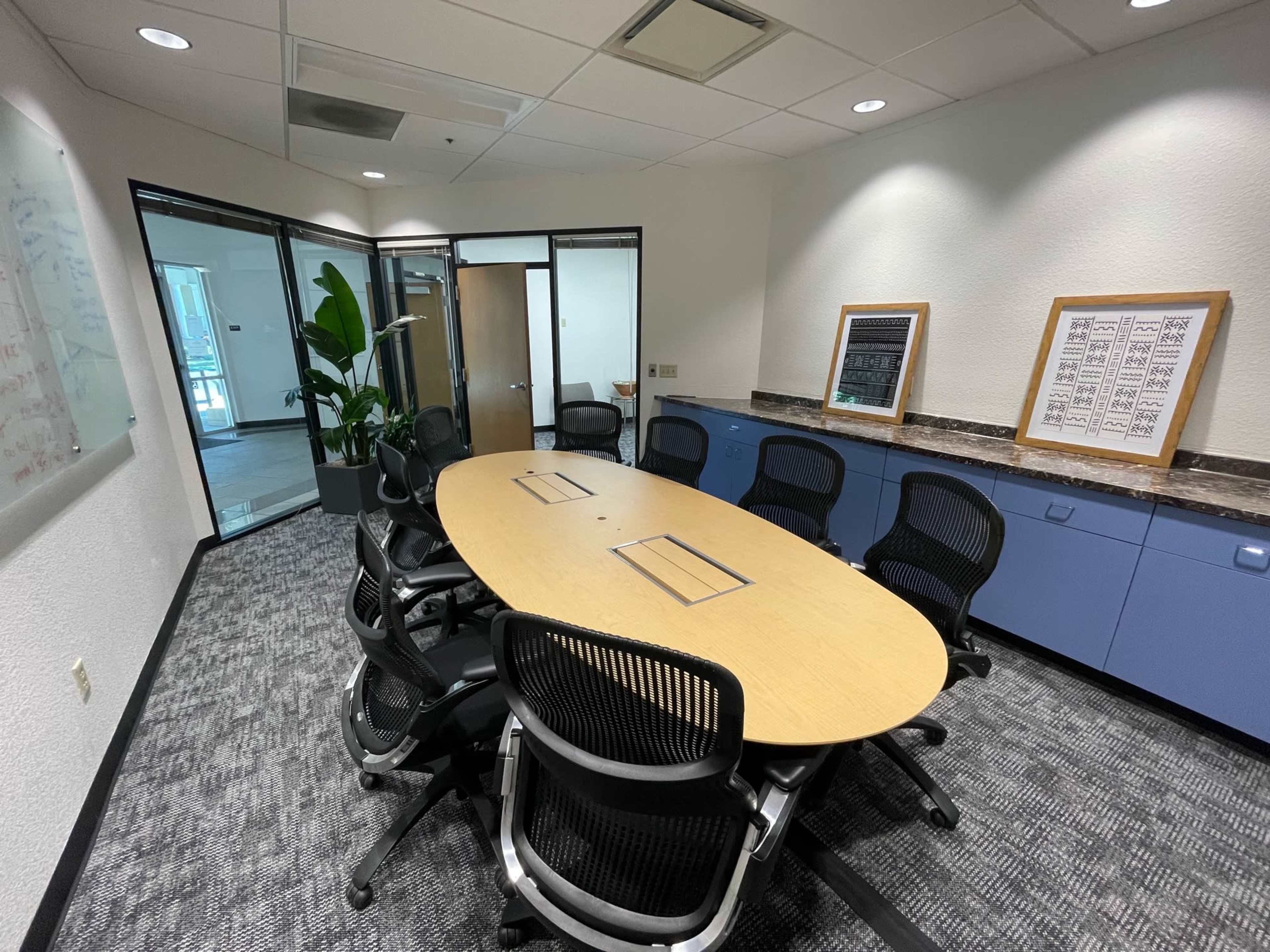 The image shows a conference room with an oval table surrounded by black chairs, adjacent to glass-walled office spaces.