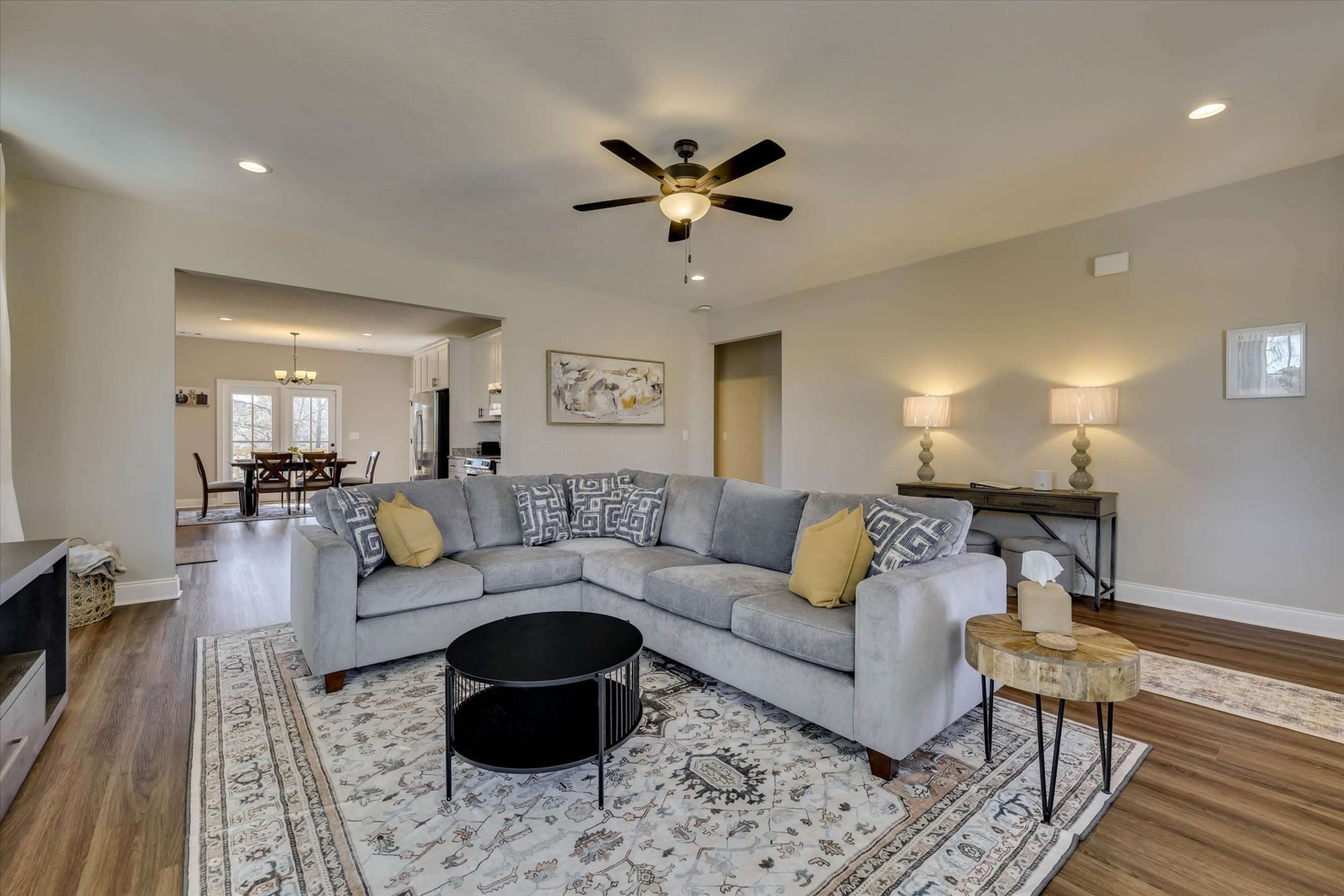 The image shows a living room with a gray sectional sofa, a round black coffee table, and a decorative rug, accompanied by a dining area visible in the background.