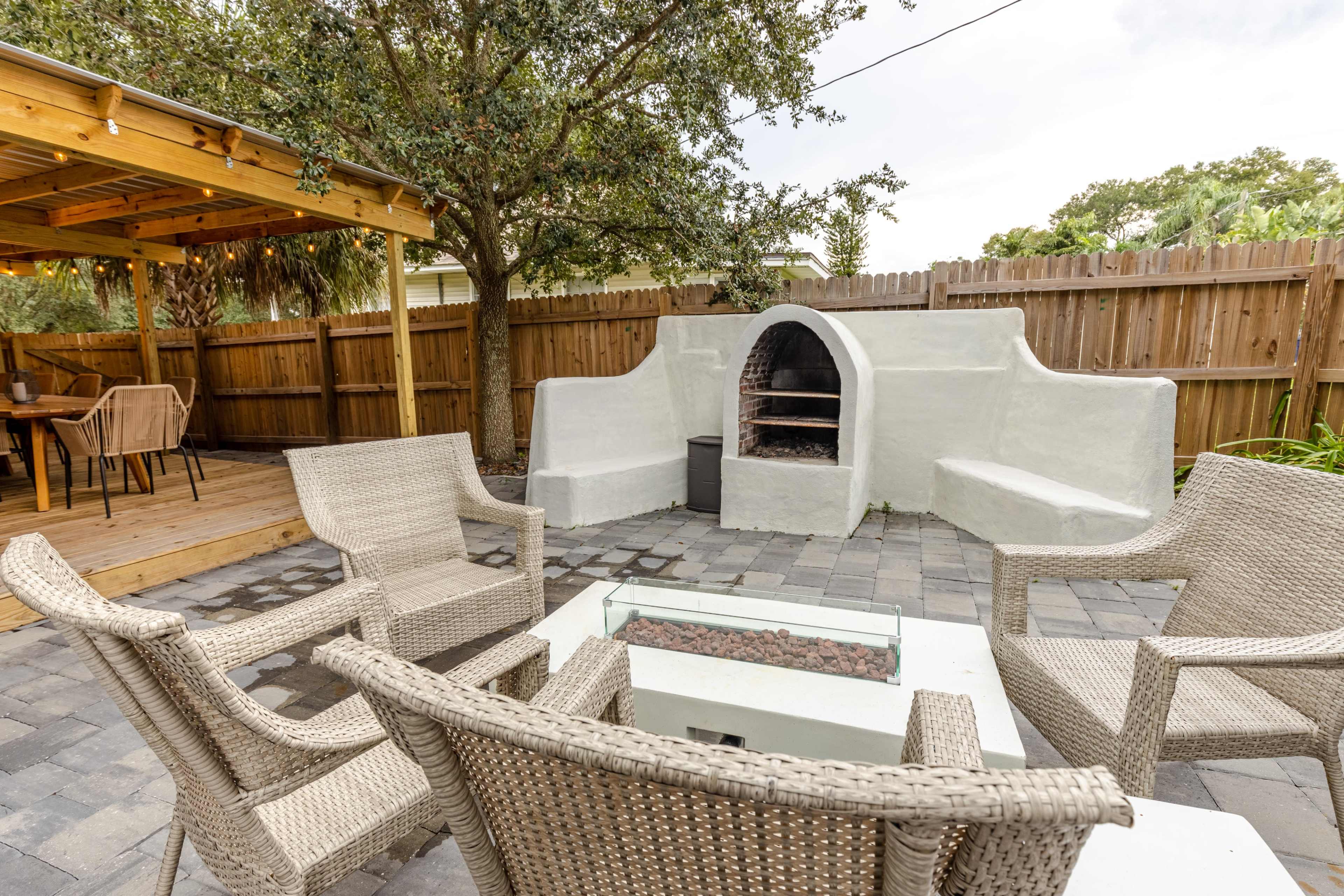A patio area with wicker chairs surrounding a fire pit, an outdoor barbecue station, and a wooden pergola nearby.