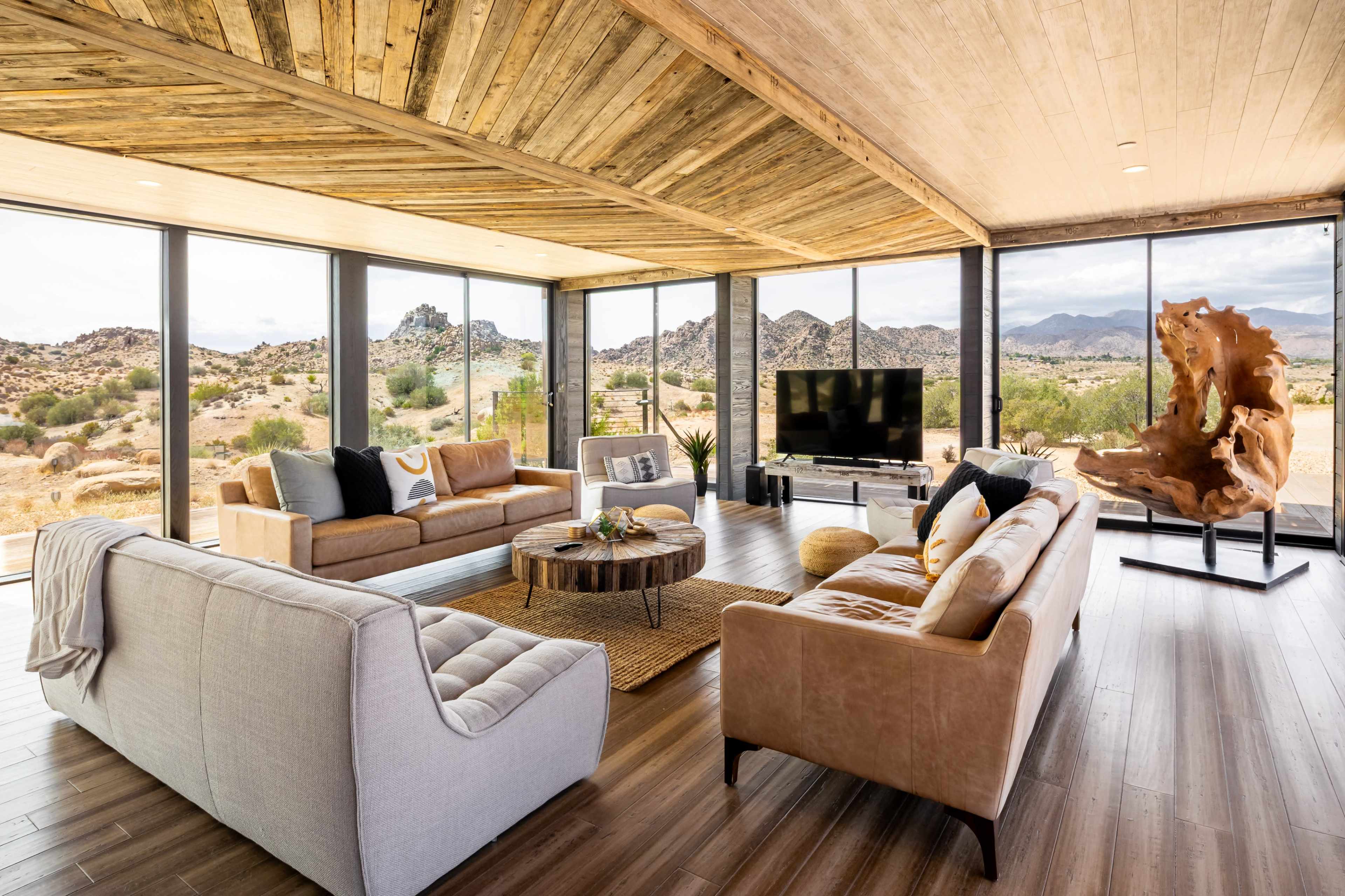 The image shows a modern living room with large glass windows overlooking a desert landscape, featuring a mix of comfortable seating, a coffee table, and a large television.