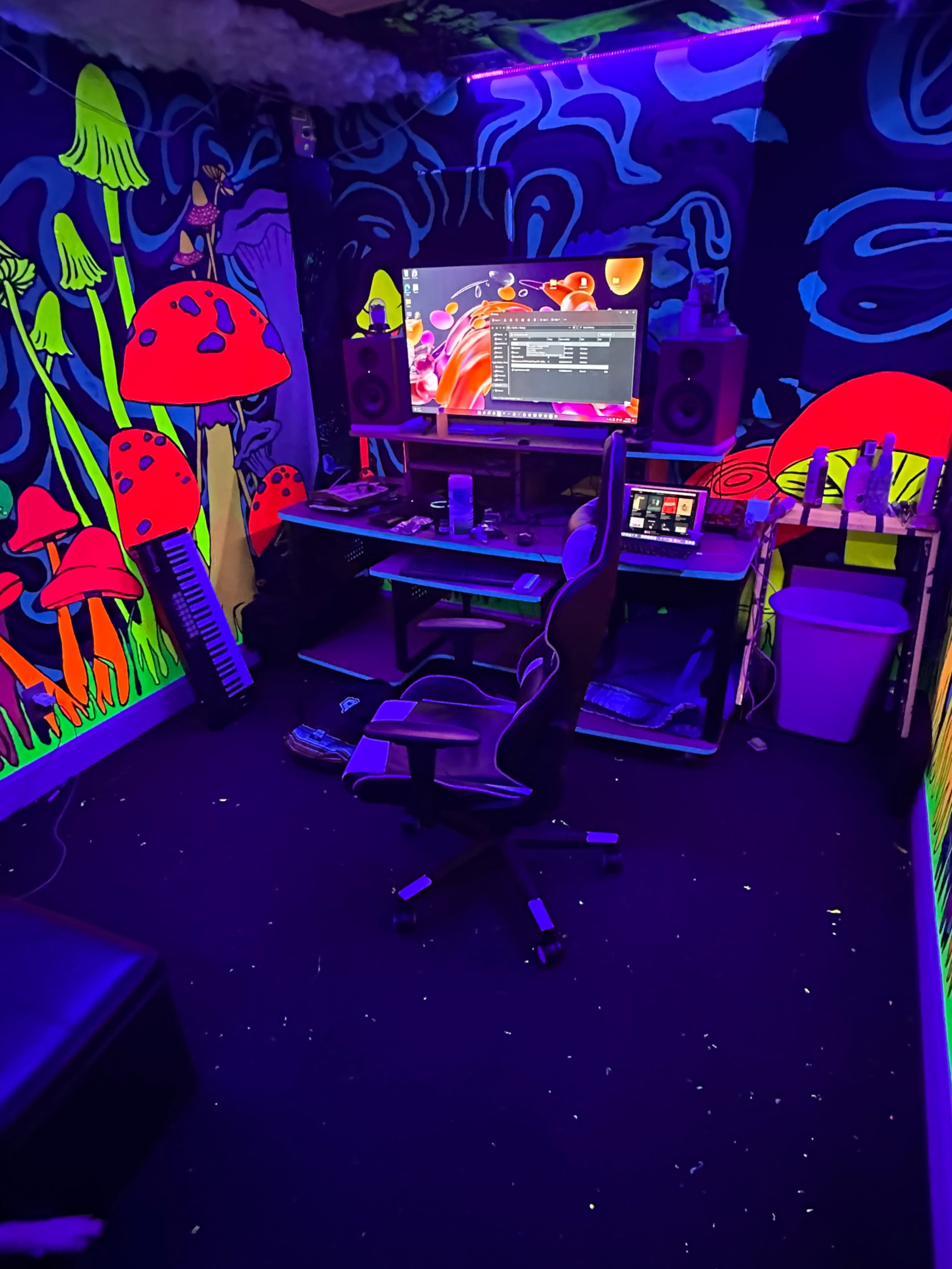 The image shows a vibrant, colorful music production studio with a computer desk, speakers, and a gaming chair, all set against a mural of bright mushrooms and a psychedelic background.