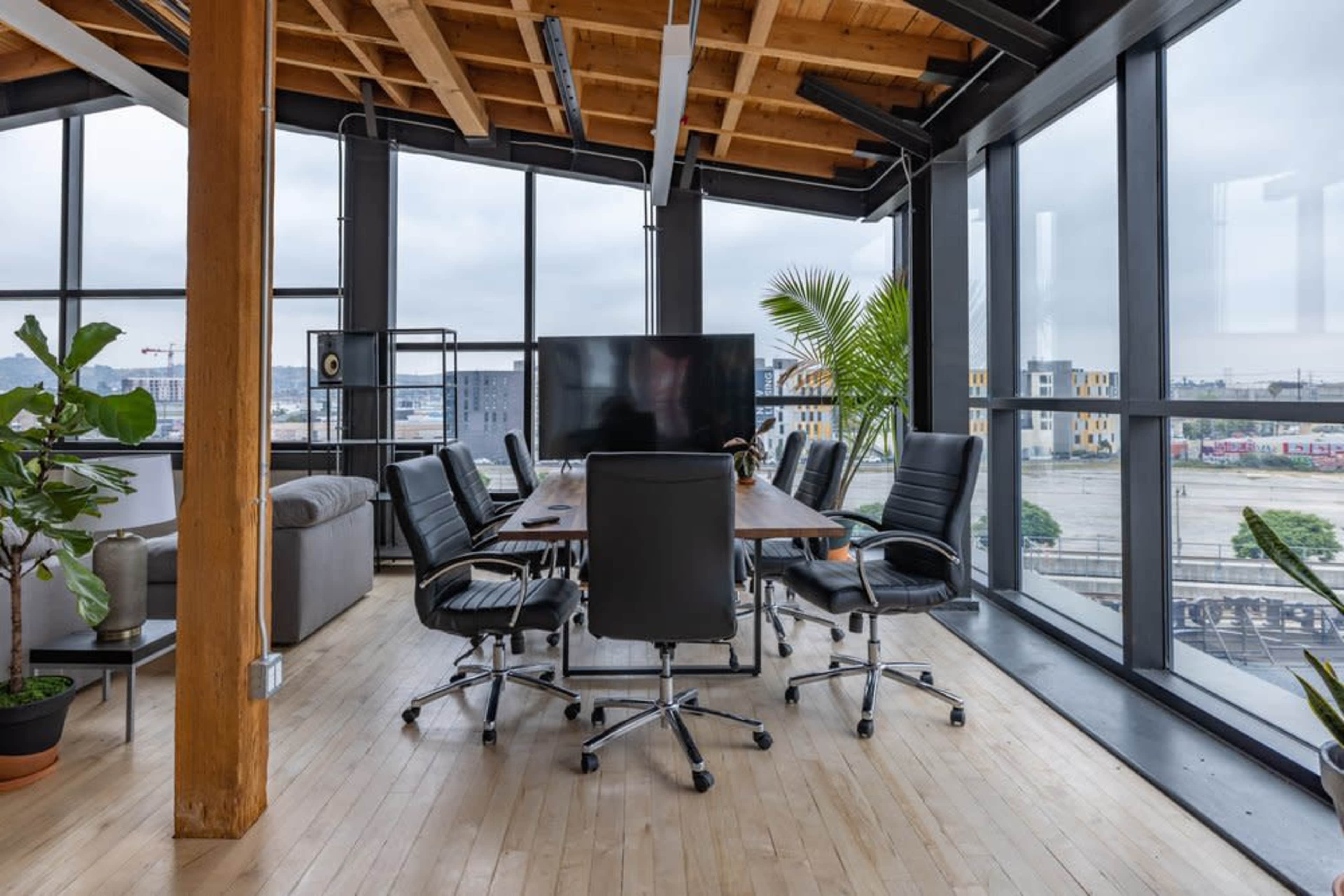 The image shows a modern conference room with large windows, a wooden table surrounded by black chairs, and greenery in the corners.