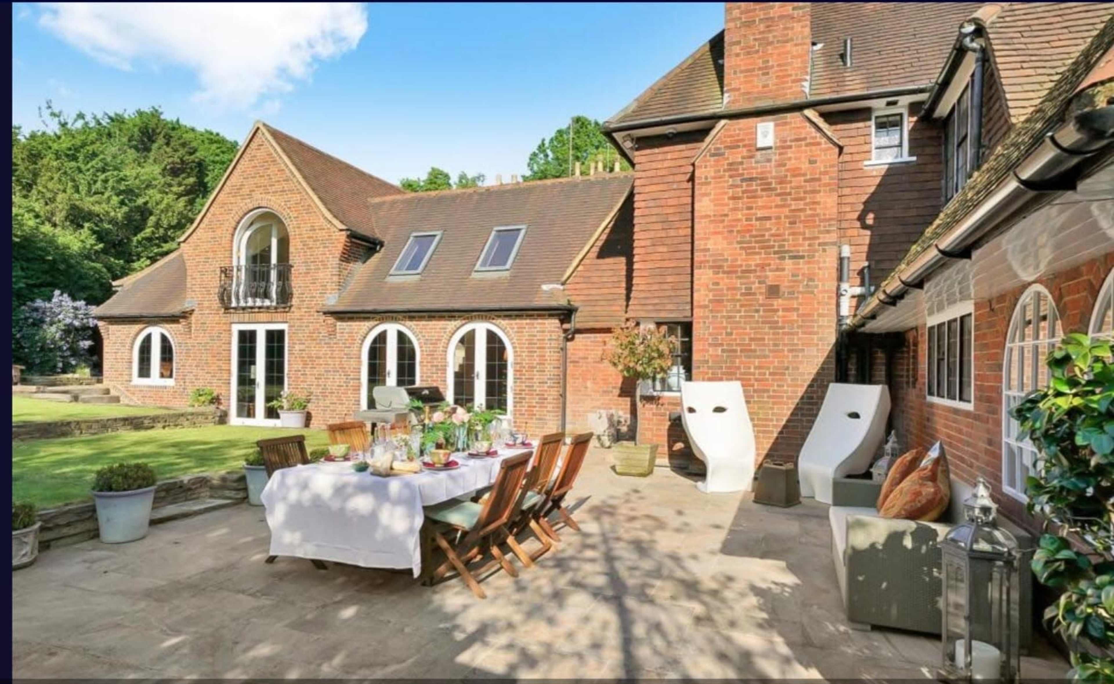 The image shows a brick house with a patio set up for dining, featuring a table with chairs and two white lounge chairs on a lawn bordered by greenery.