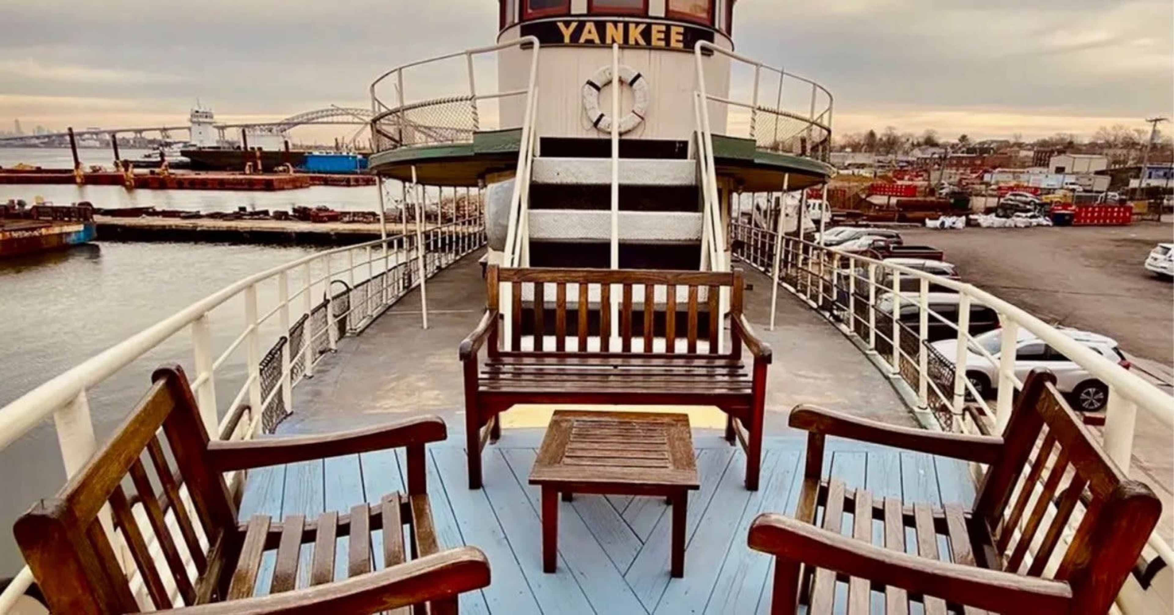 The image shows a view of the deck of a boat named "Yankee" featuring two wooden chairs and a small table, with a harbor and industrial area in the background.