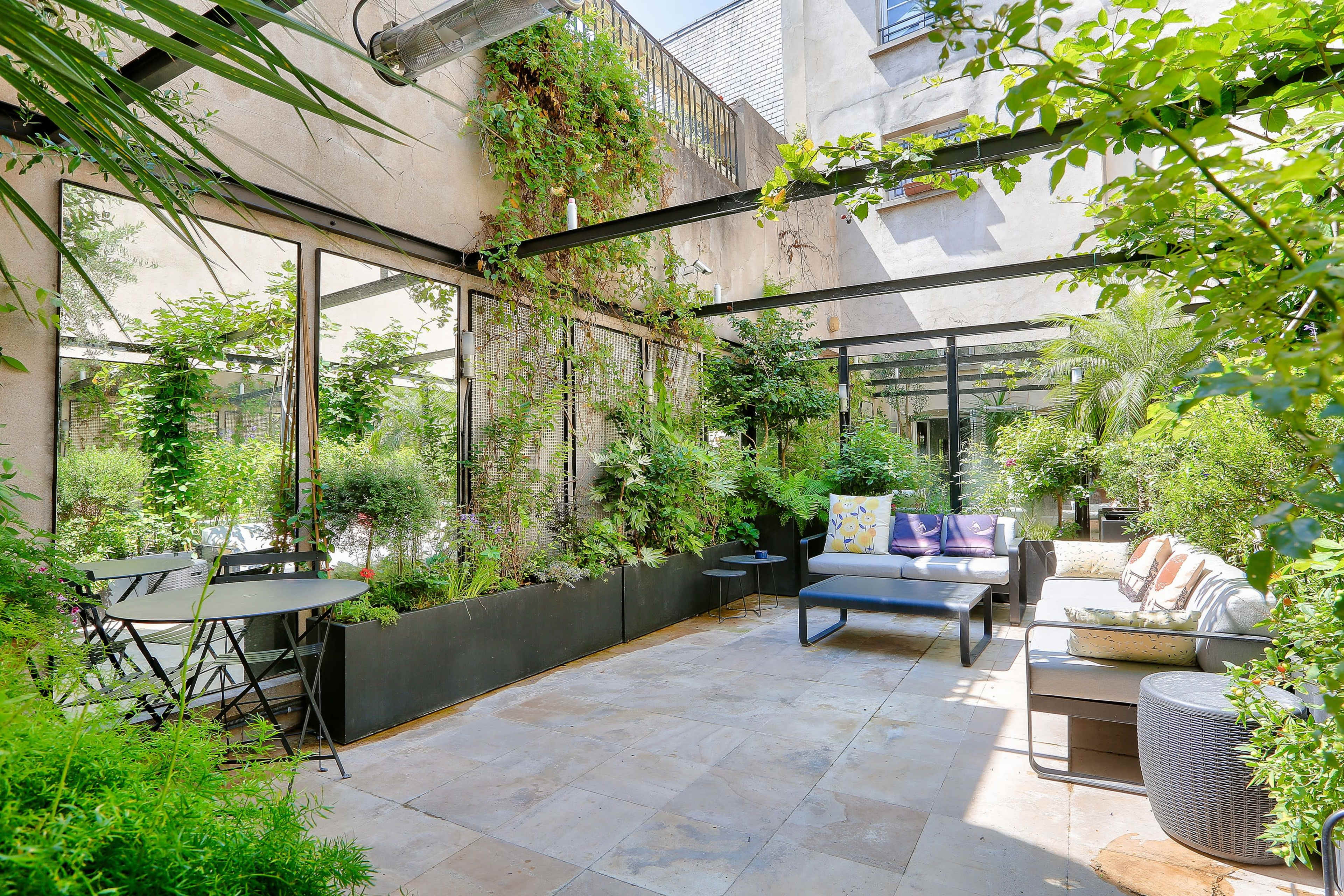 The image shows a well-lit, lush garden terrace featuring a seating area with a sofa, a bistro table, and various plants surrounding the space.