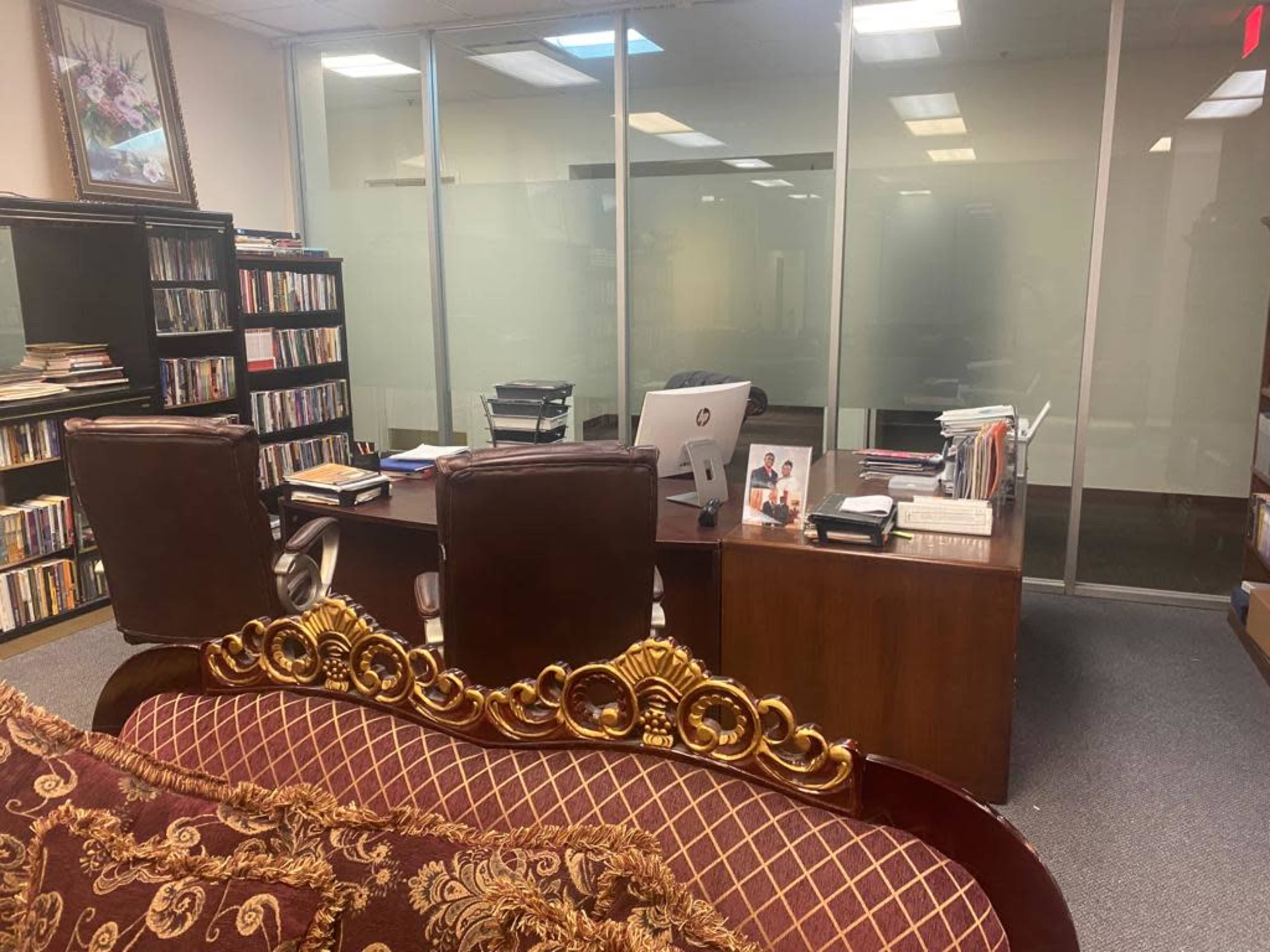 An office space features a wooden desk with two chairs, a bookshelf filled with DVDs, and a couch in the foreground.