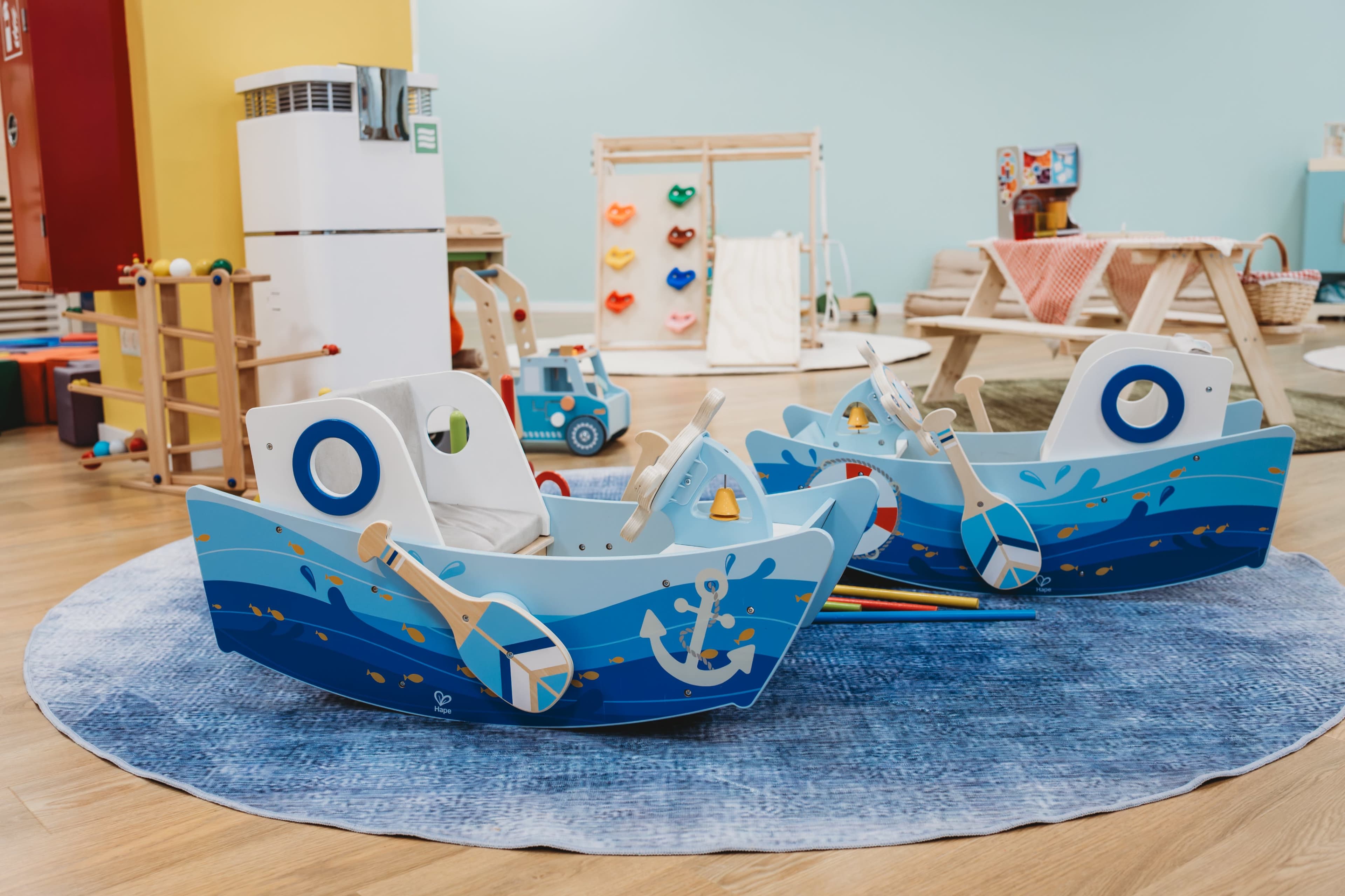 The image shows a colorful children’s play area featuring two wooden boat-shaped play structures on a round blue rug, surrounded by various toys and furniture.