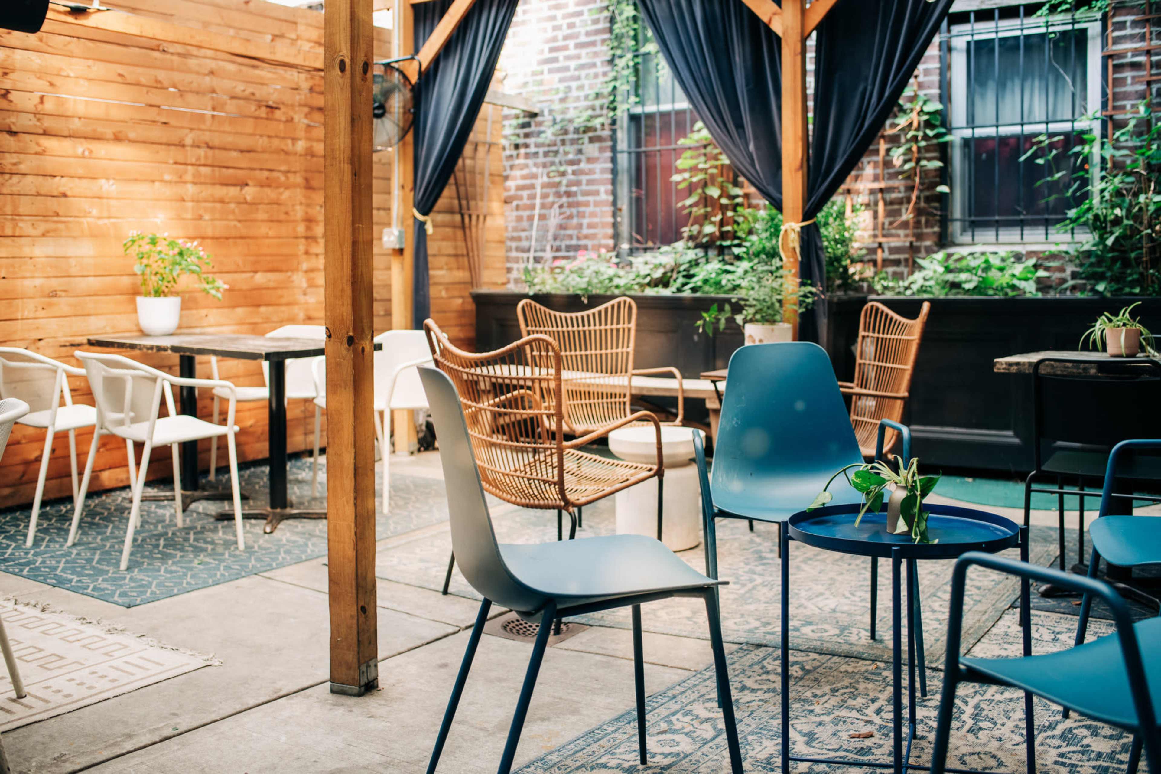 The image shows a cozy outdoor seating area with wooden walls and neatly arranged tables and chairs, surrounded by greenery.