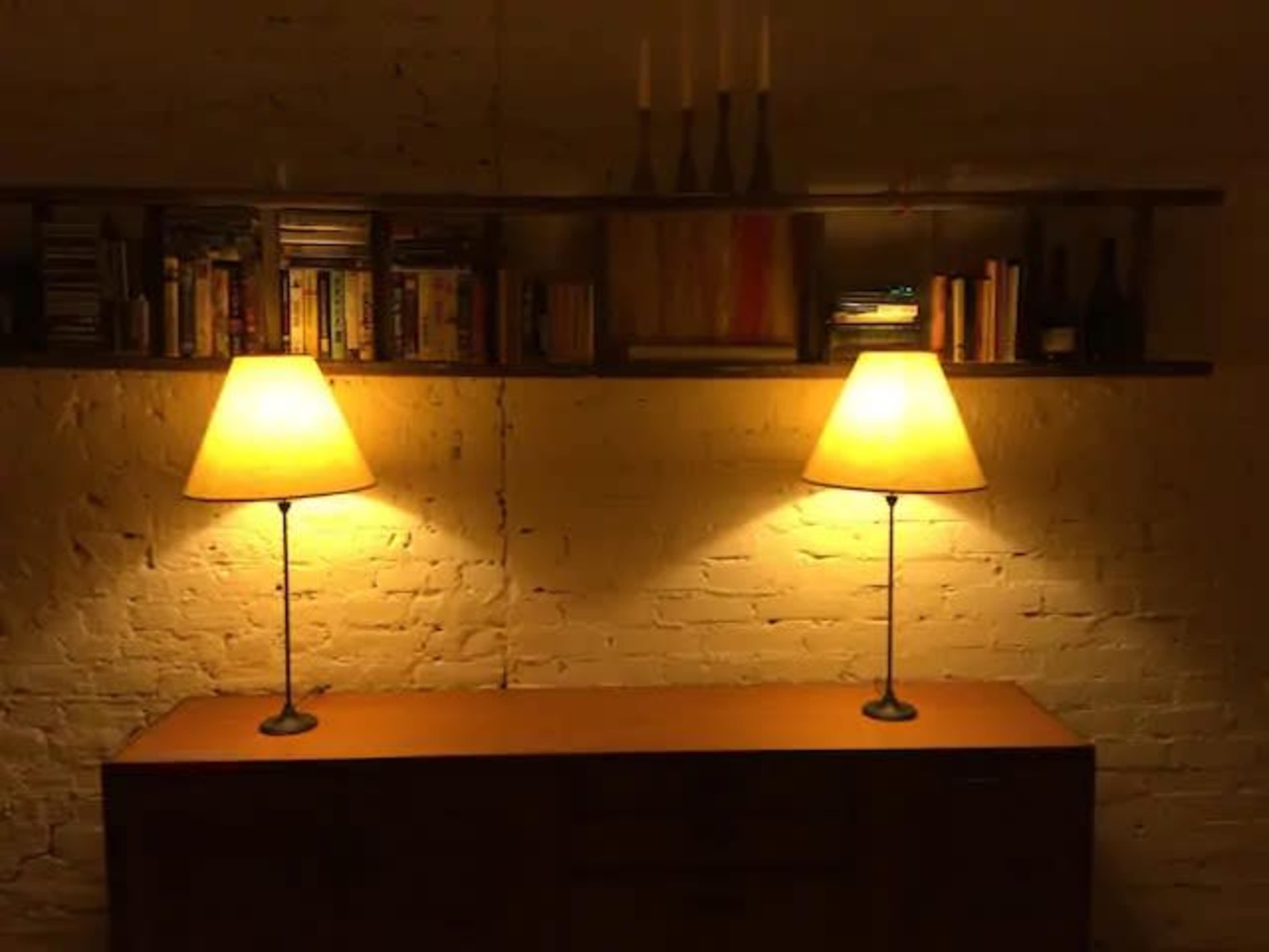 The image shows a dimly lit room with two lamps casting light on a wooden sideboard beneath a shelf filled with books and decorative items.