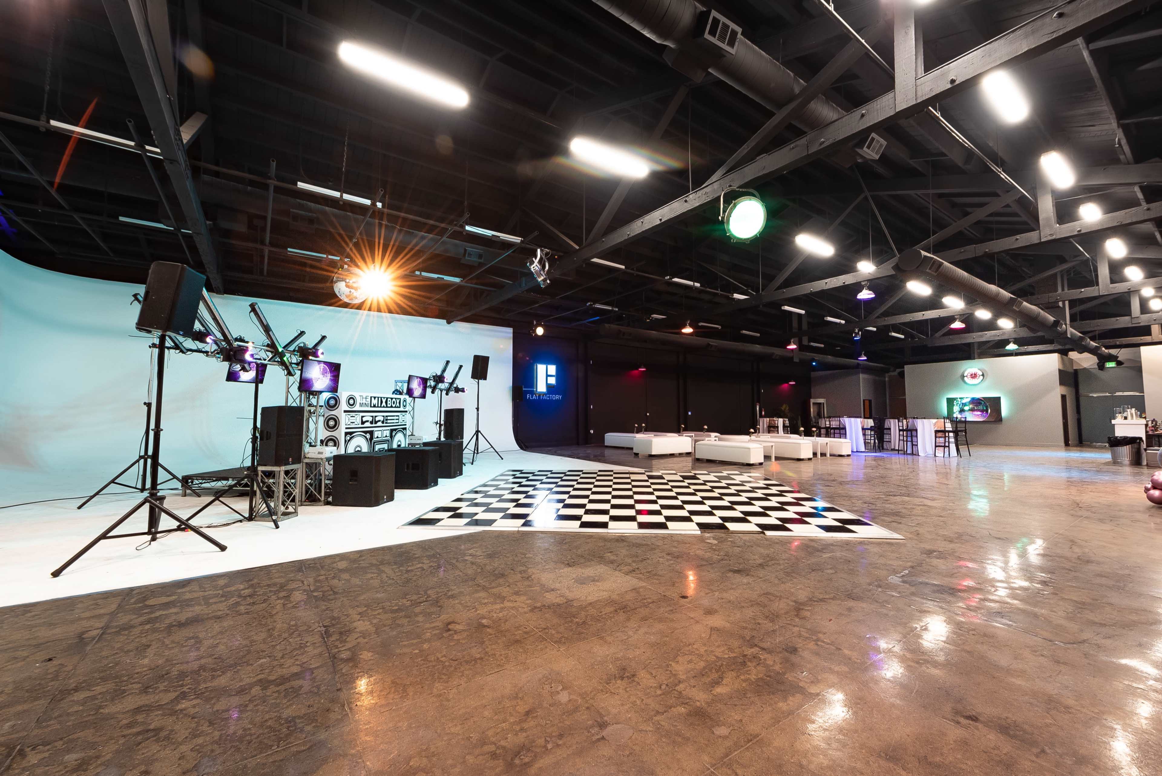 The image shows a spacious event venue featuring a DJ setup, a checkered dance floor, white seating areas, and colorful lighting.