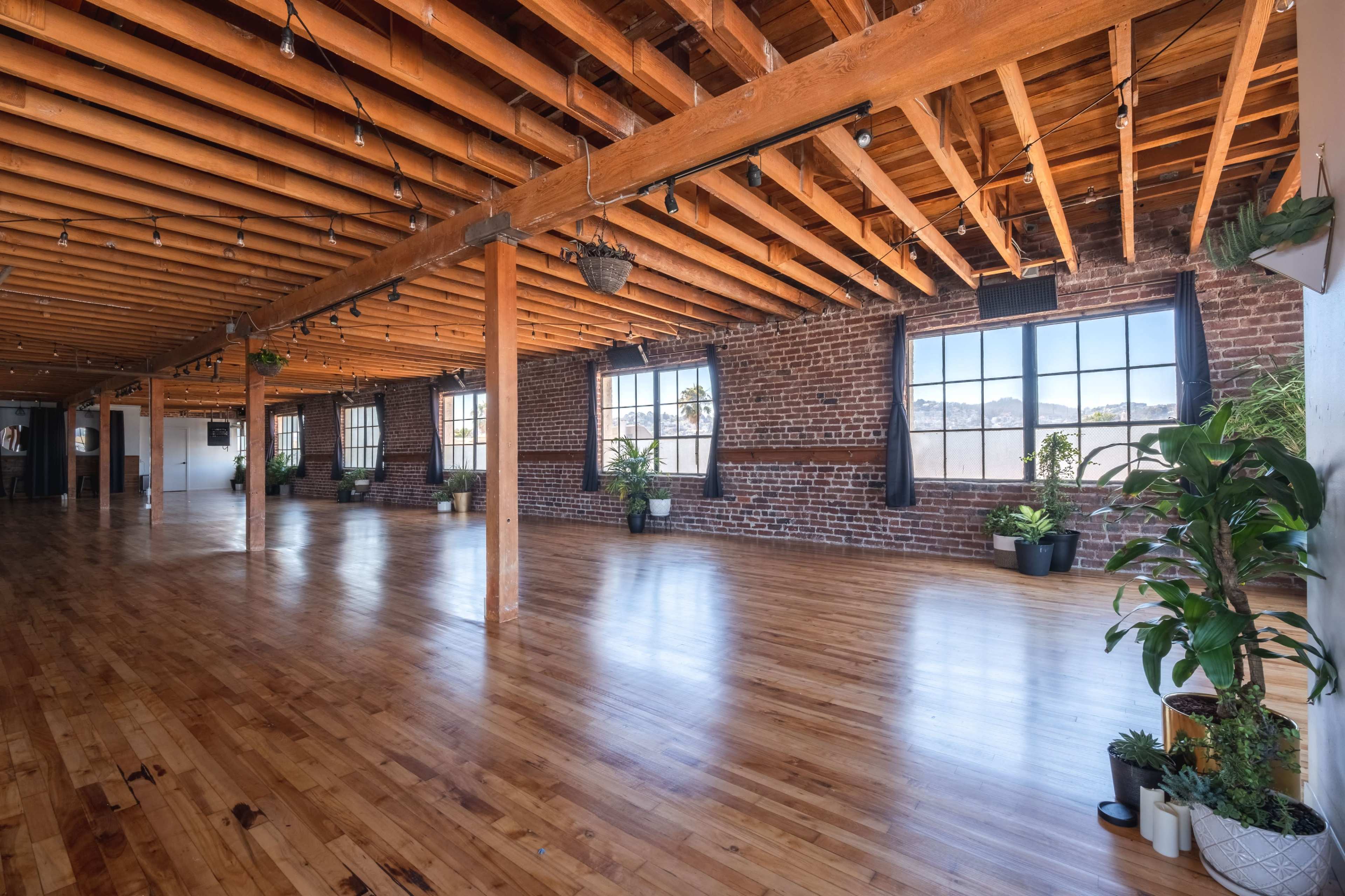 A spacious indoor area featuring exposed wooden beams, brick walls, and large windows with plants positioned around the room.