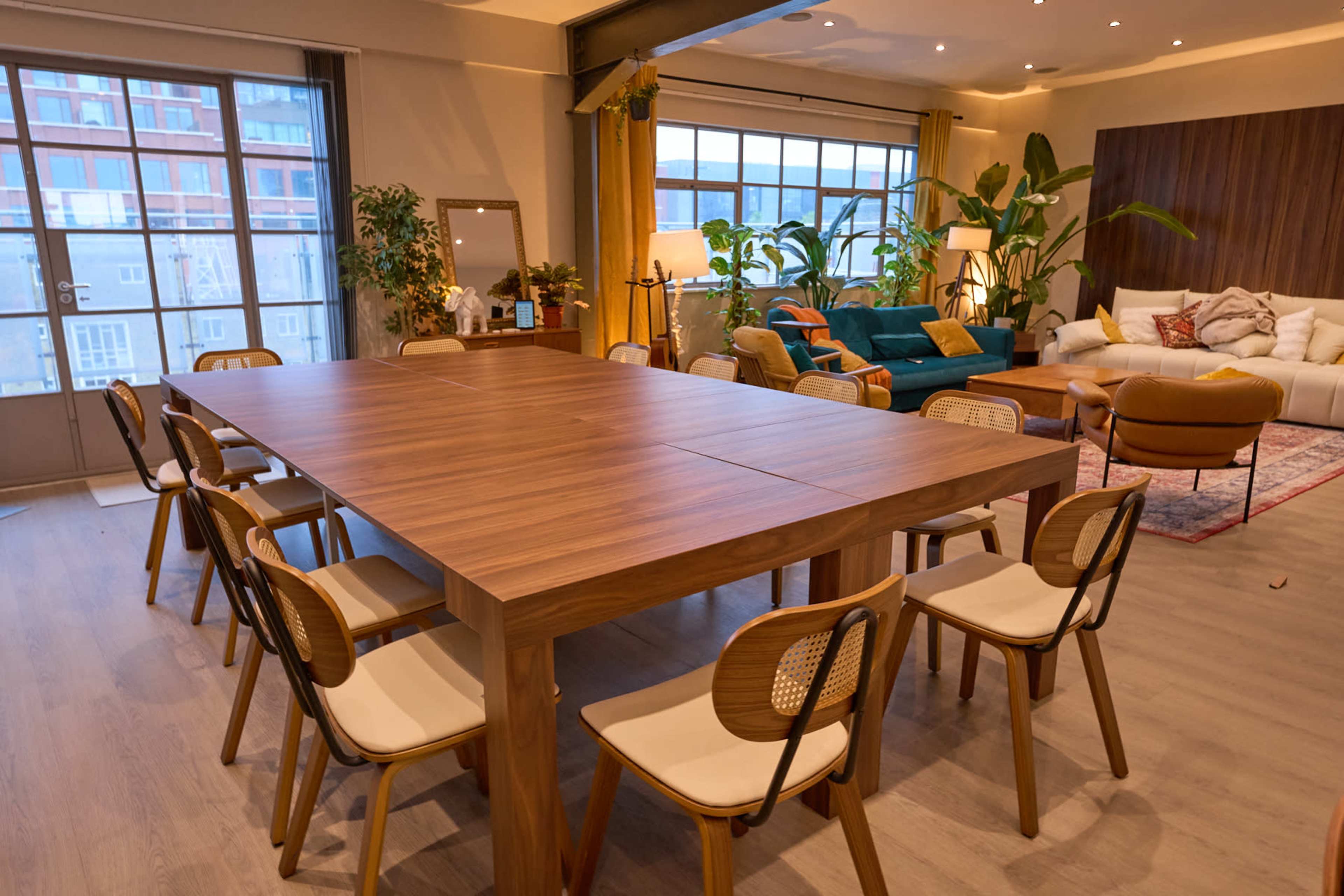 A spacious dining area features a large wooden table surrounded by white and wooden chairs, with a cozy living room area in the background that includes a green couch and various plants.
