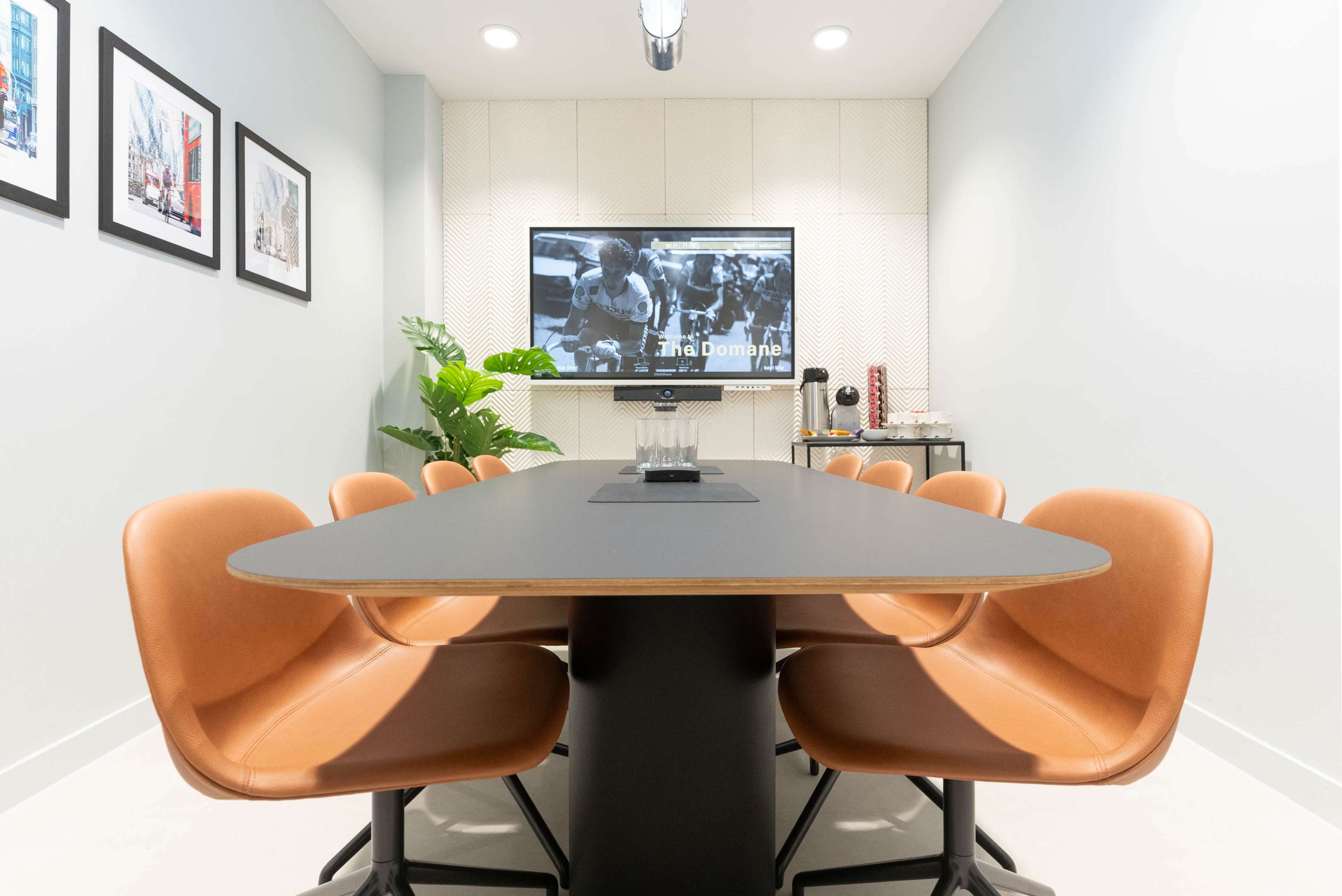 A modern conference room features a long black table surrounded by brown chairs, with a television displaying black-and-white imagery on one wall.