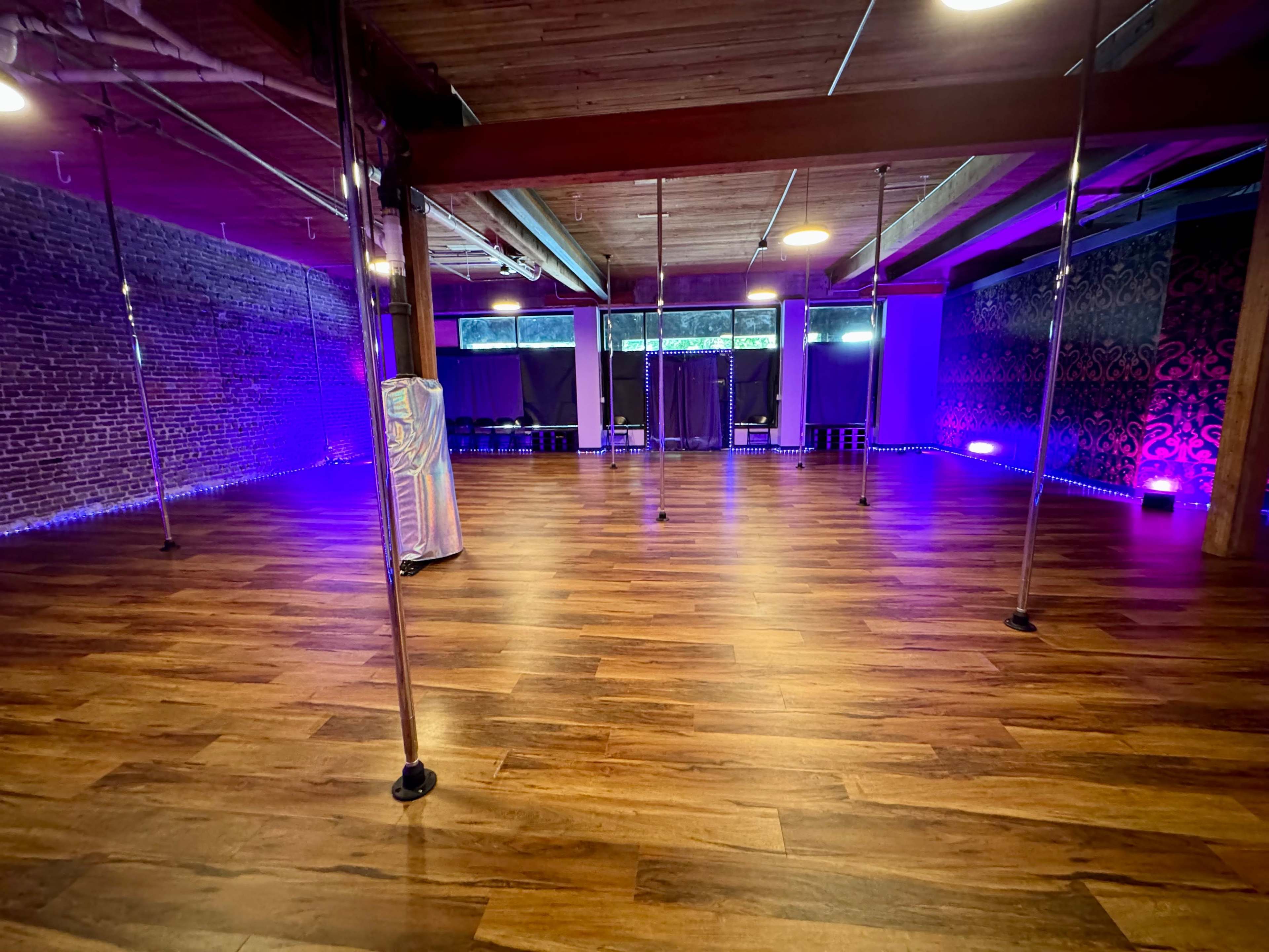 A spacious dance studio with polished wooden floors, several vertical poles, and purple lighting along the walls.