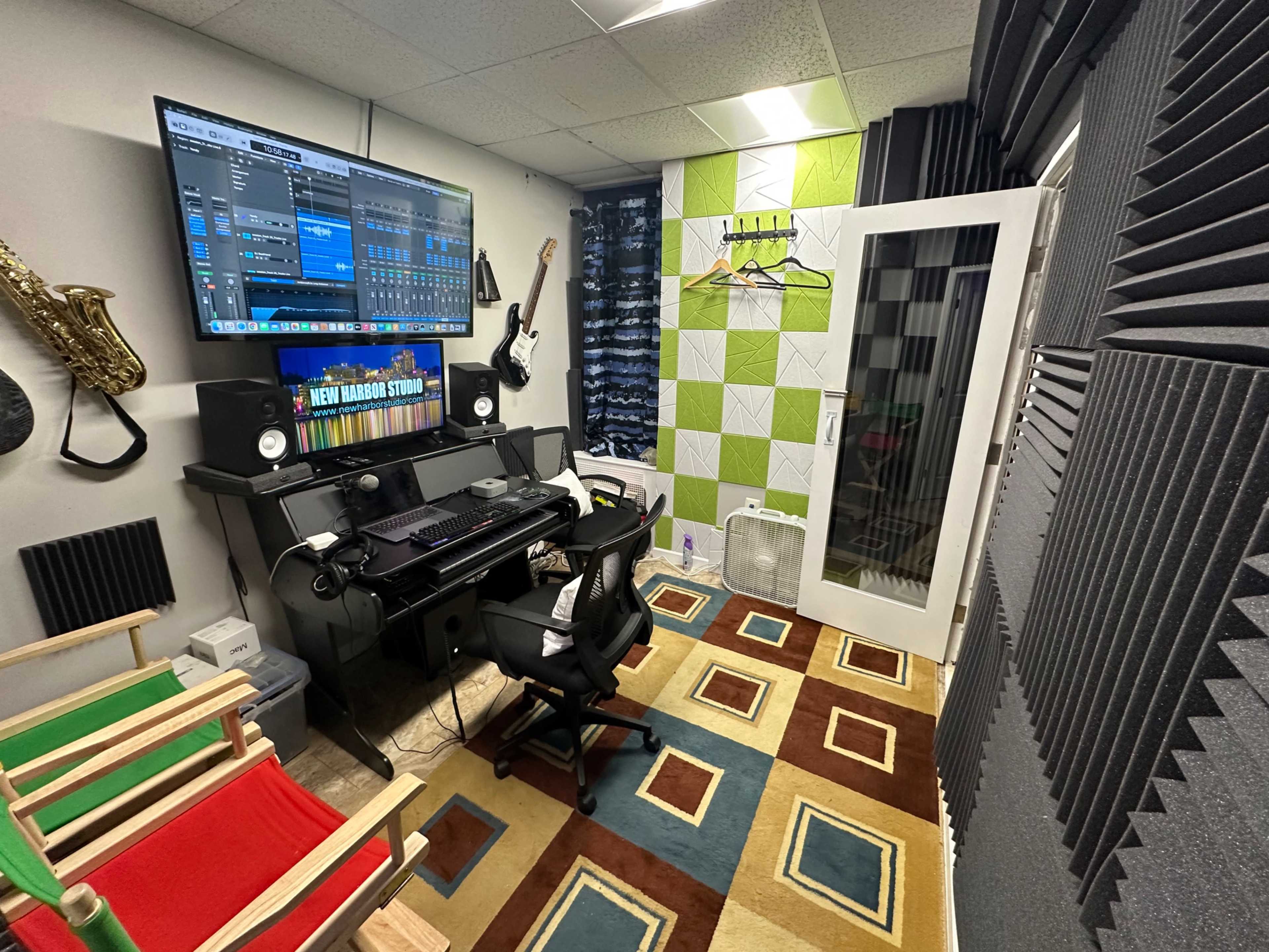The image shows a soundproof music studio with a desk, computer screen displaying audio software, and various musical instruments on the walls, alongside colorful accents and acoustic panels.