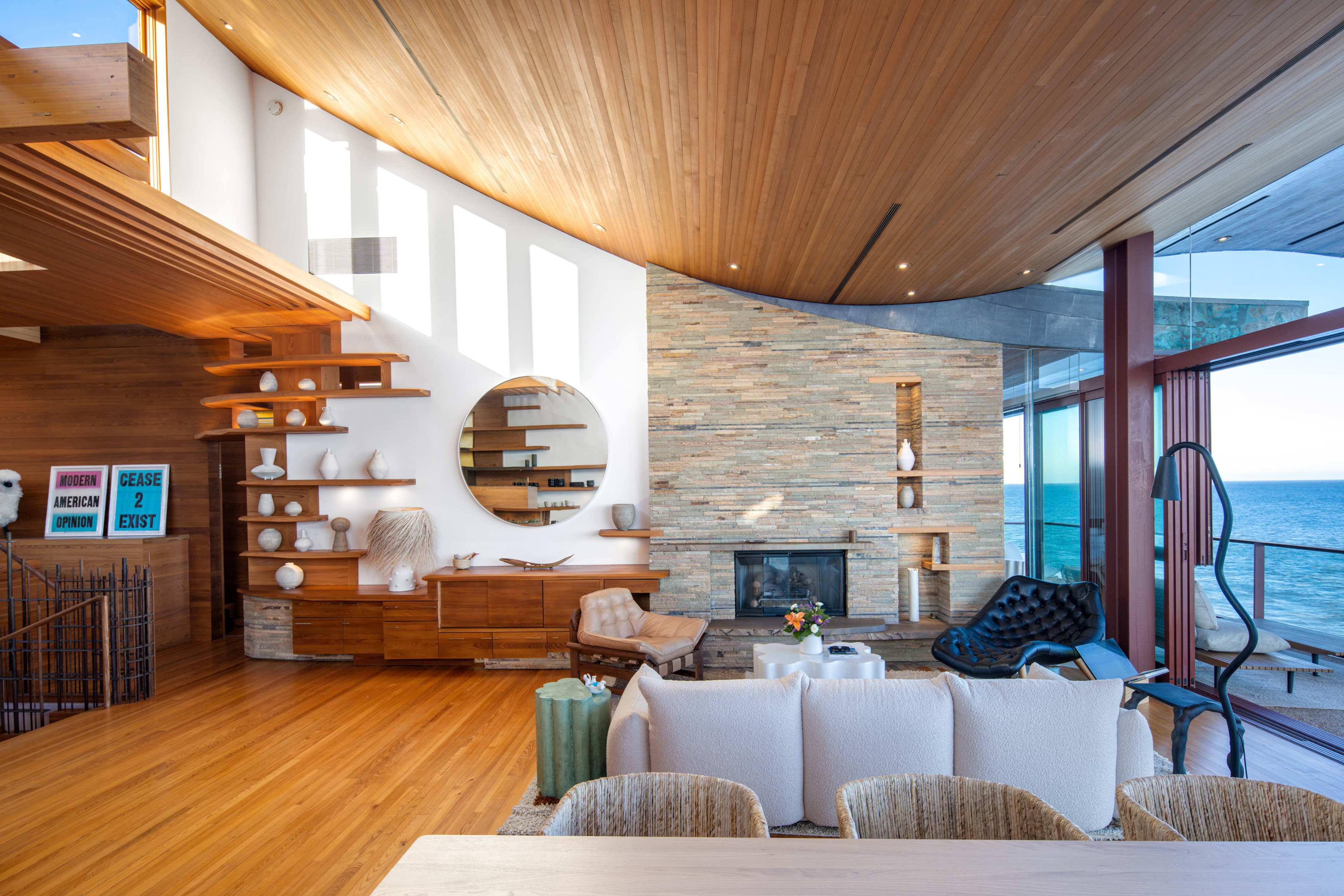 A modern living room features a curved wooden ceiling, a stone accent wall with a fireplace, and large windows overlooking the ocean.