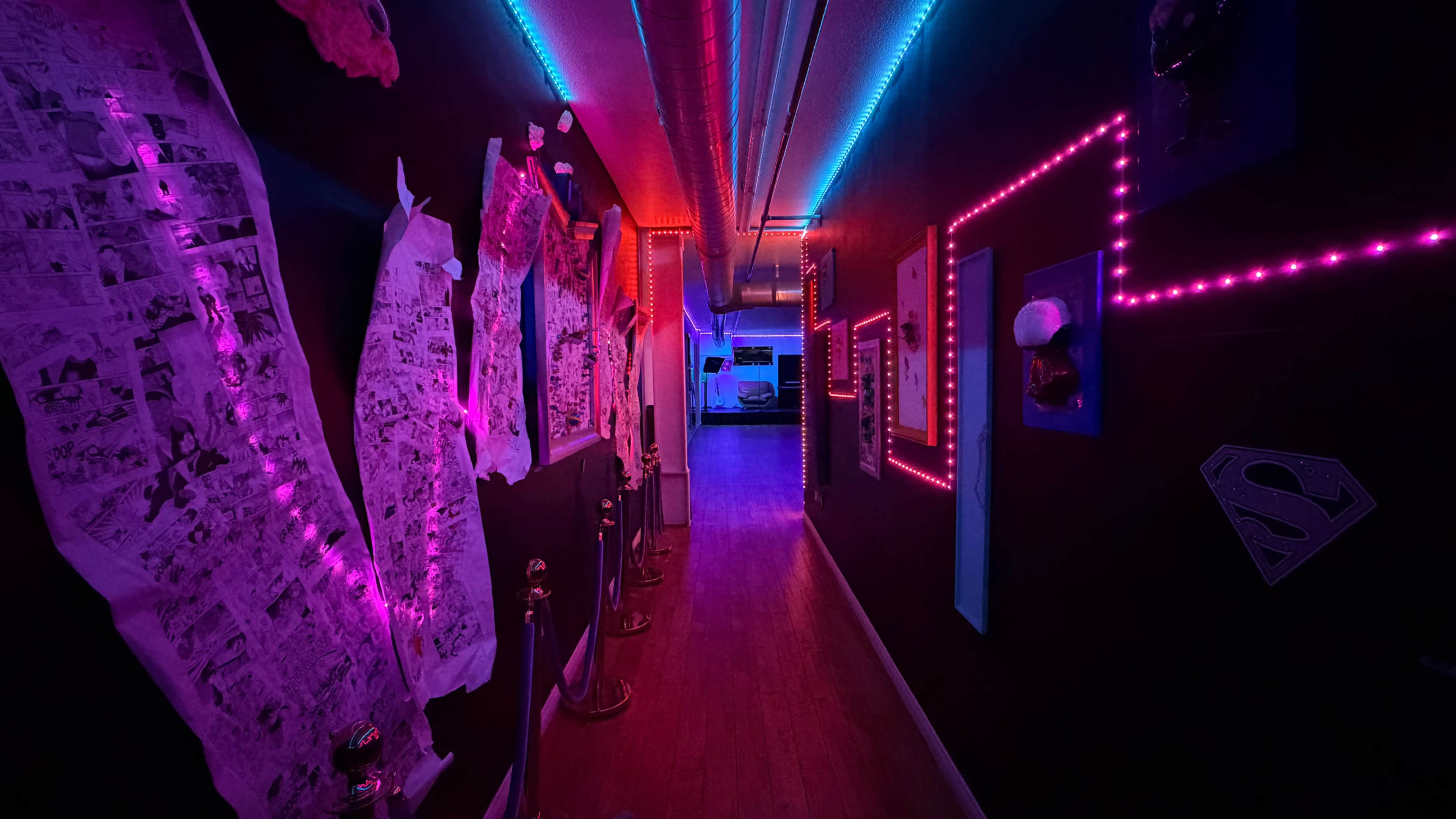 The image depicts a dimly lit hallway with colorful neon lights and framed artworks lining the walls.