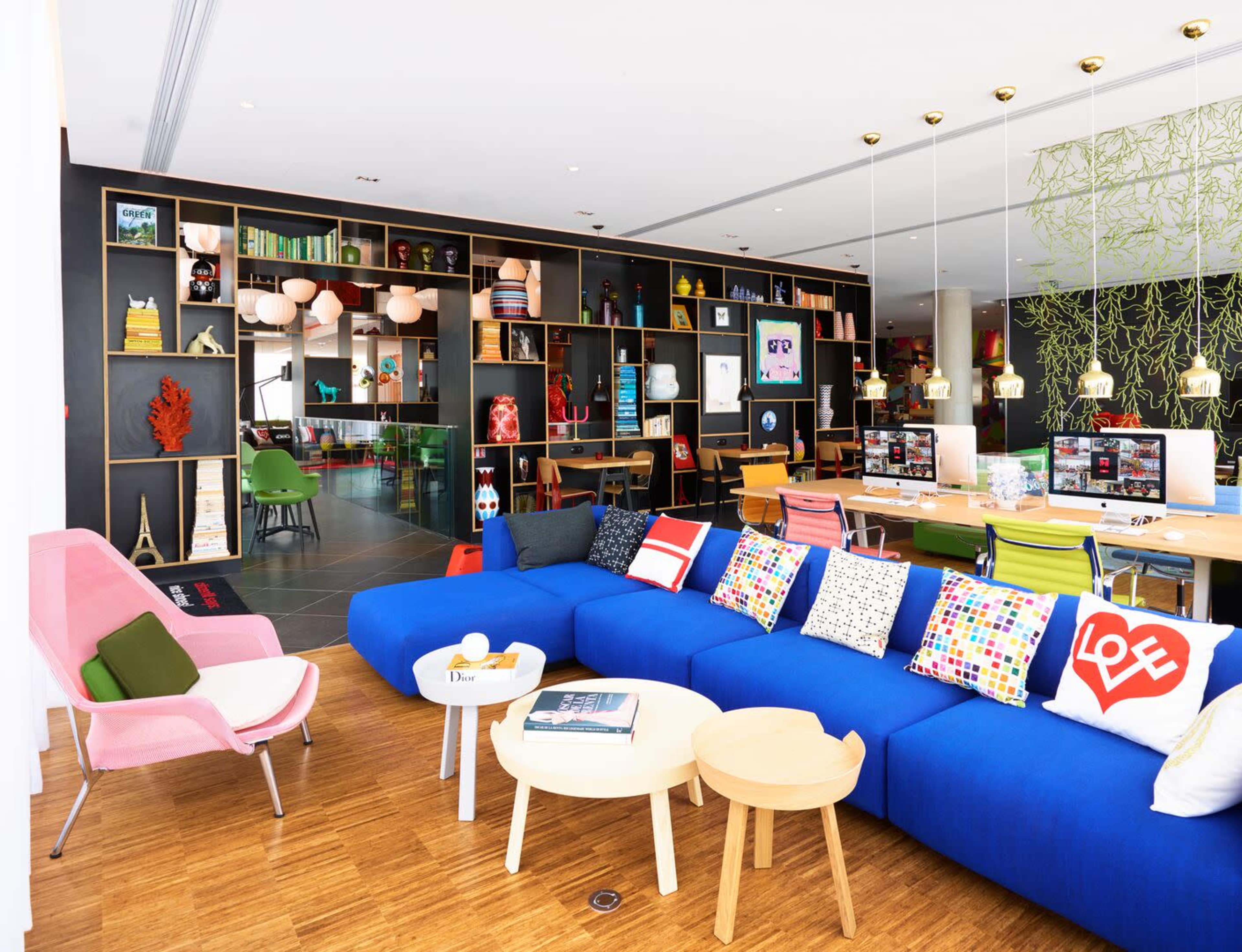 A modern interior featuring a blue sofa with decorative pillows, wooden tables, and a colorful bookshelf filled with various objects and art pieces.