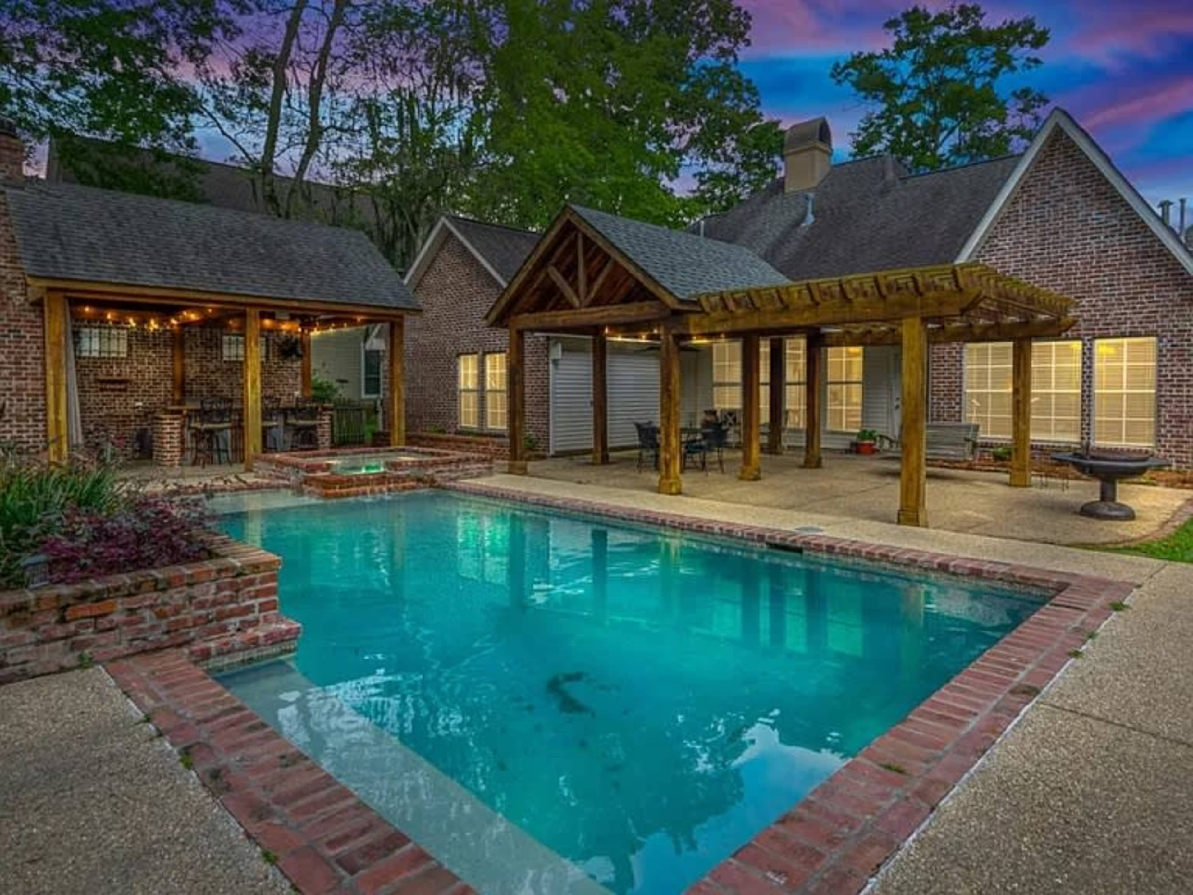 The image shows a backyard with a swimming pool, brick patio, and a covered seating area surrounded by trees and a house.
