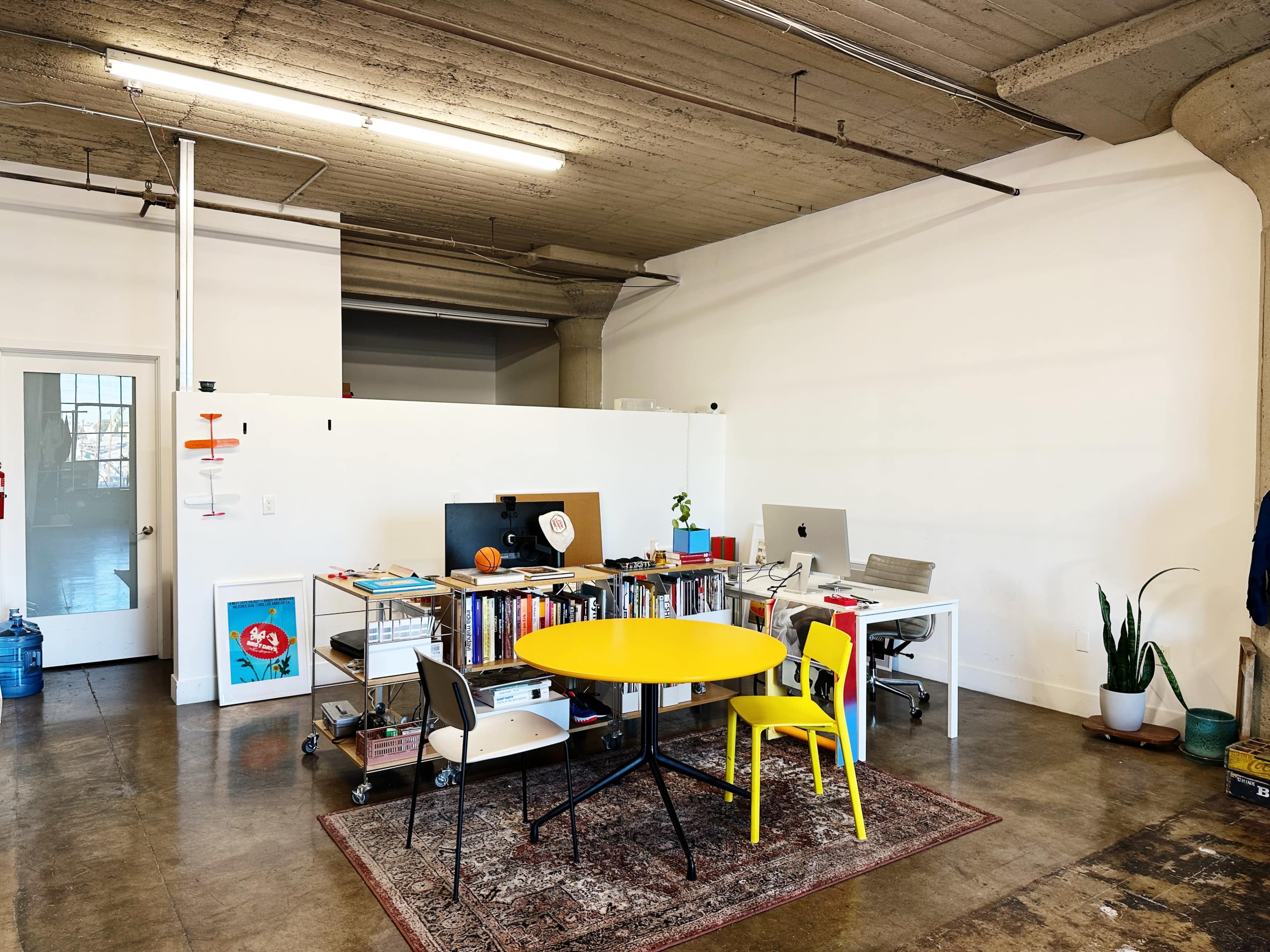 The image shows a minimalistic office space featuring a yellow table with two chairs, a desk with a computer, shelves filled with books, and a plant in the corner.
