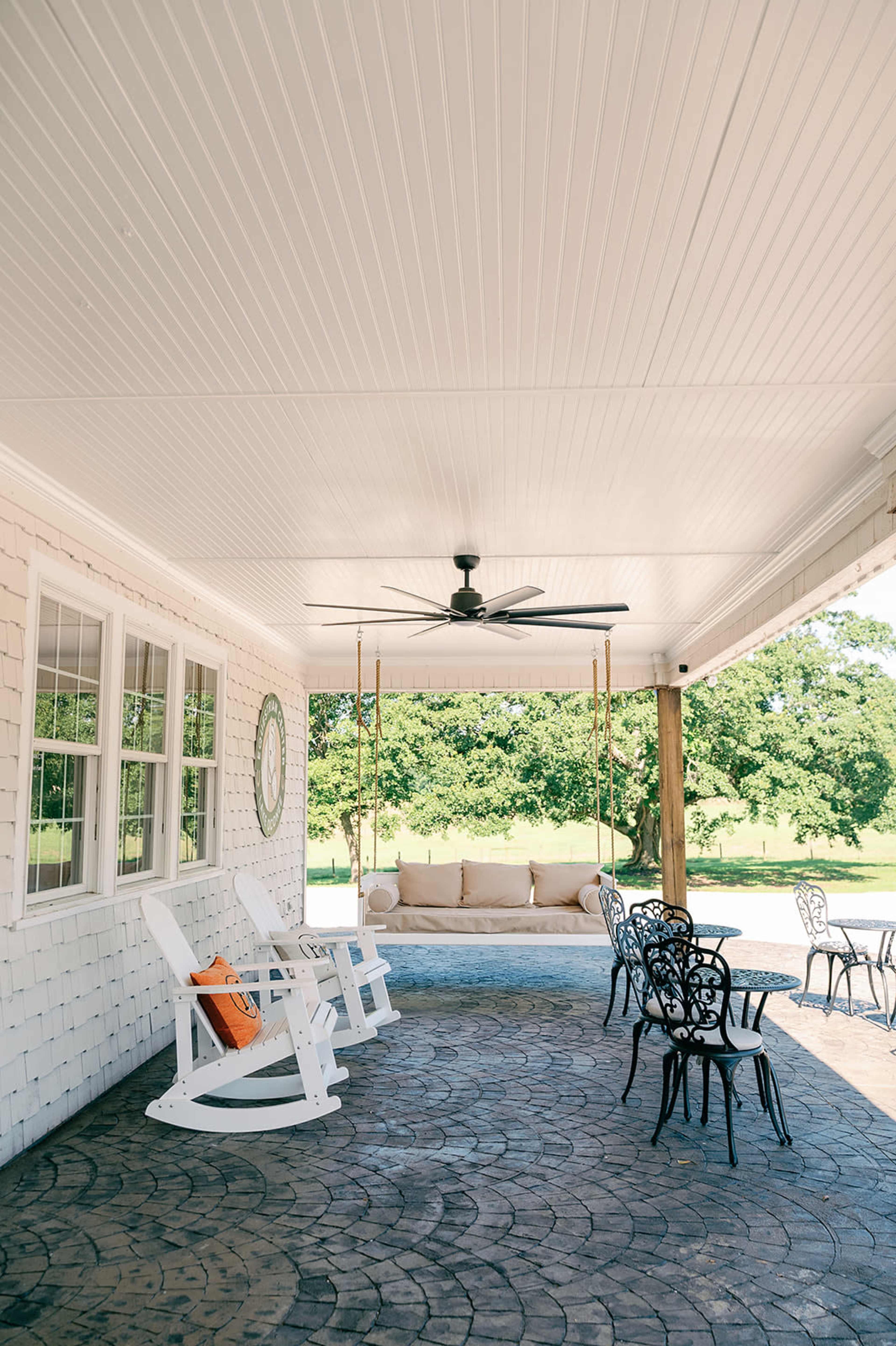 The image shows a patio with white rocking chairs, a sofa, and black metal chairs, all beneath a ceiling fan and a white ceiling.