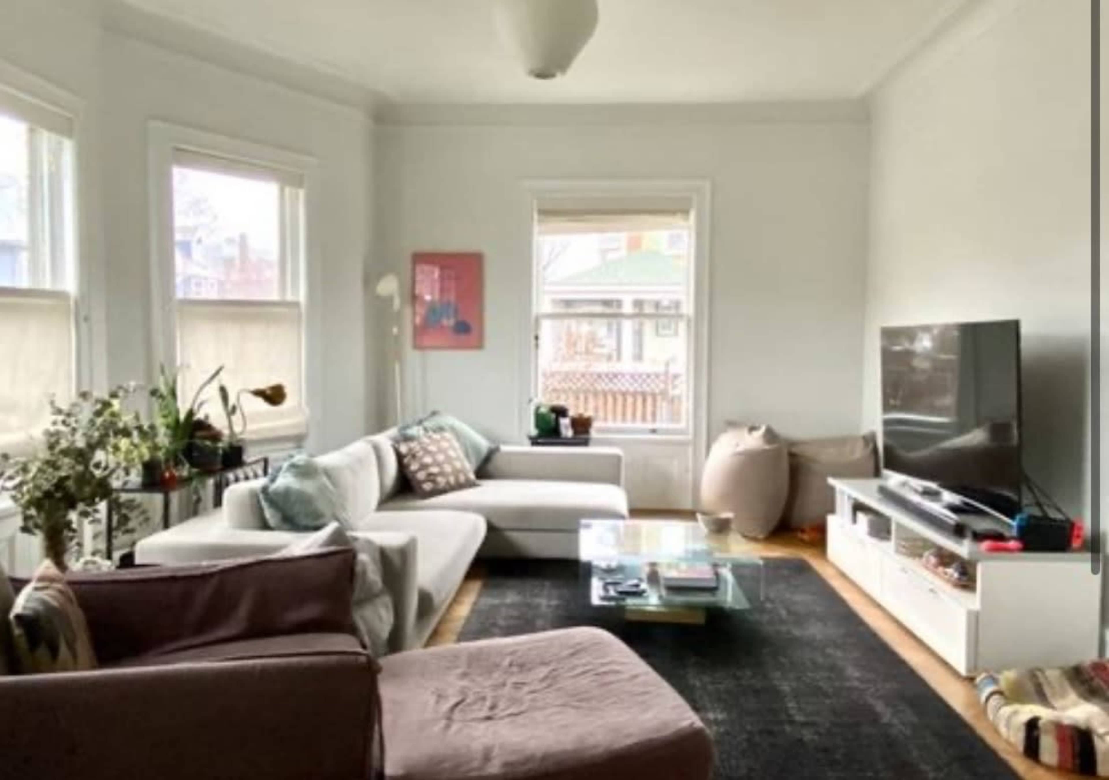 The image shows a living room with a light-colored sofa, a glass coffee table, a large television, and various plants near the windows.