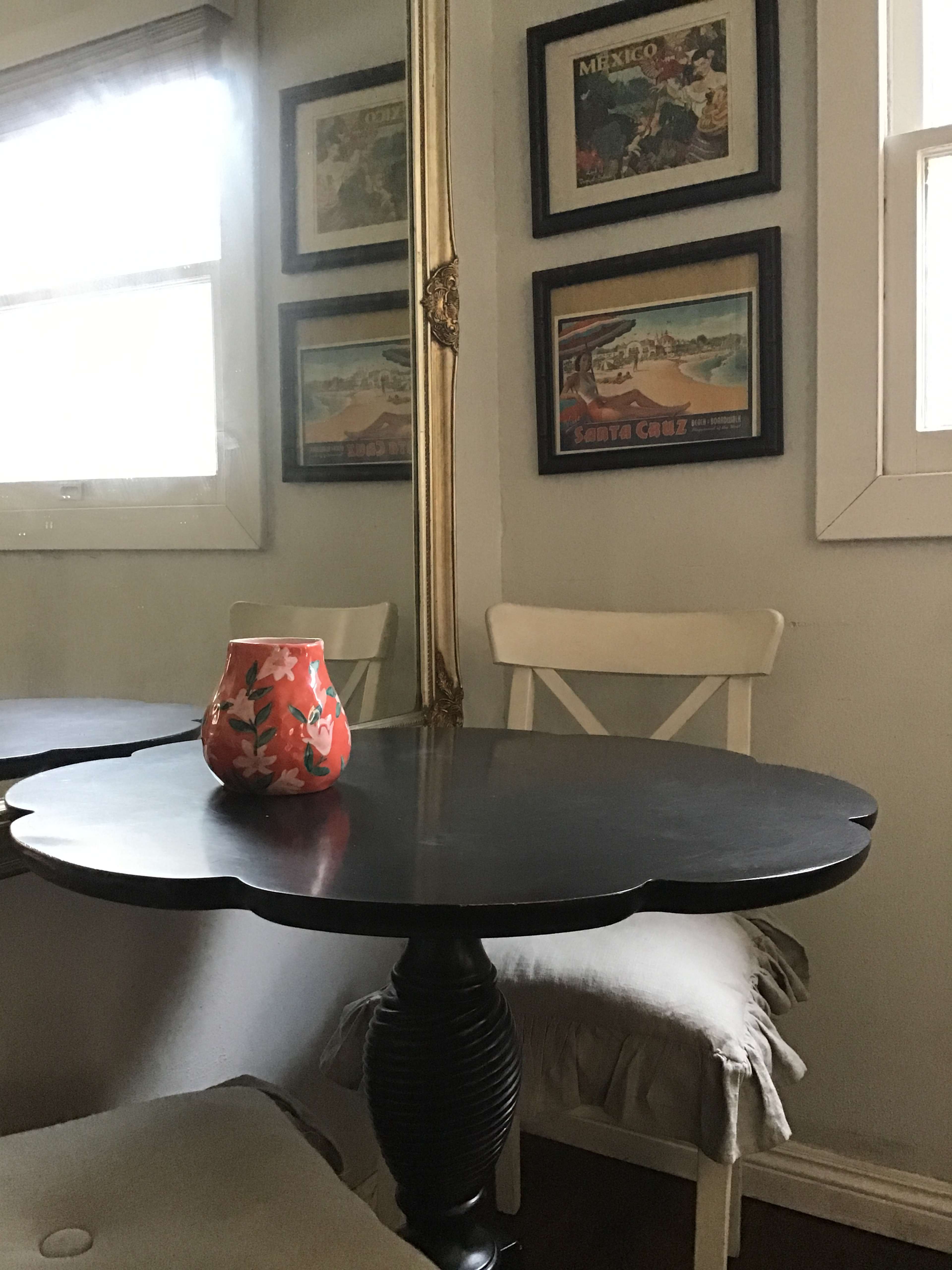 A small black table with a decorative edge and a colorful vase sits in a room with framed vintage travel posters on the walls.