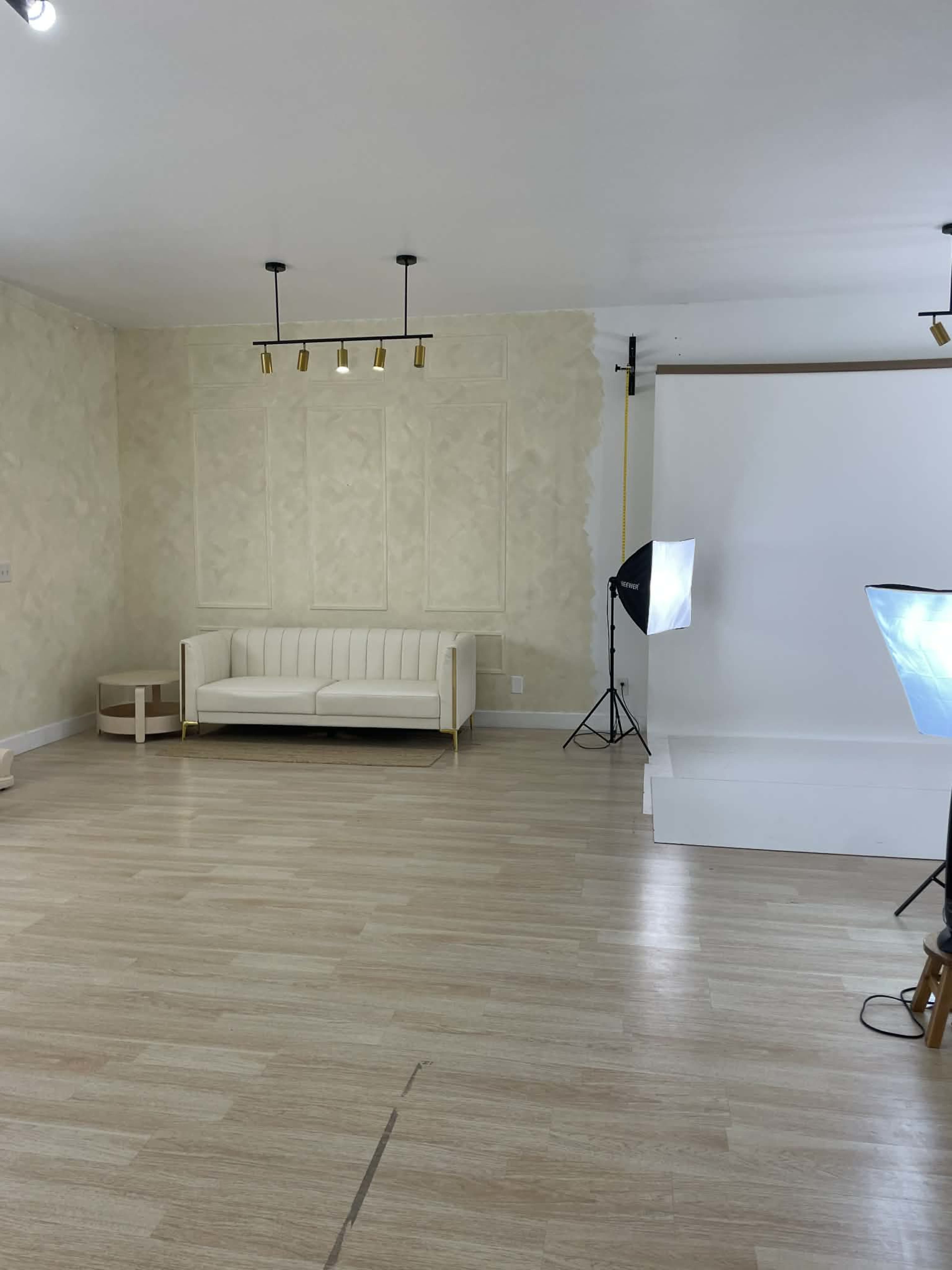 The image shows a well-lit studio space featuring a white couch, a small table, and a backdrop stand with lighting equipment.