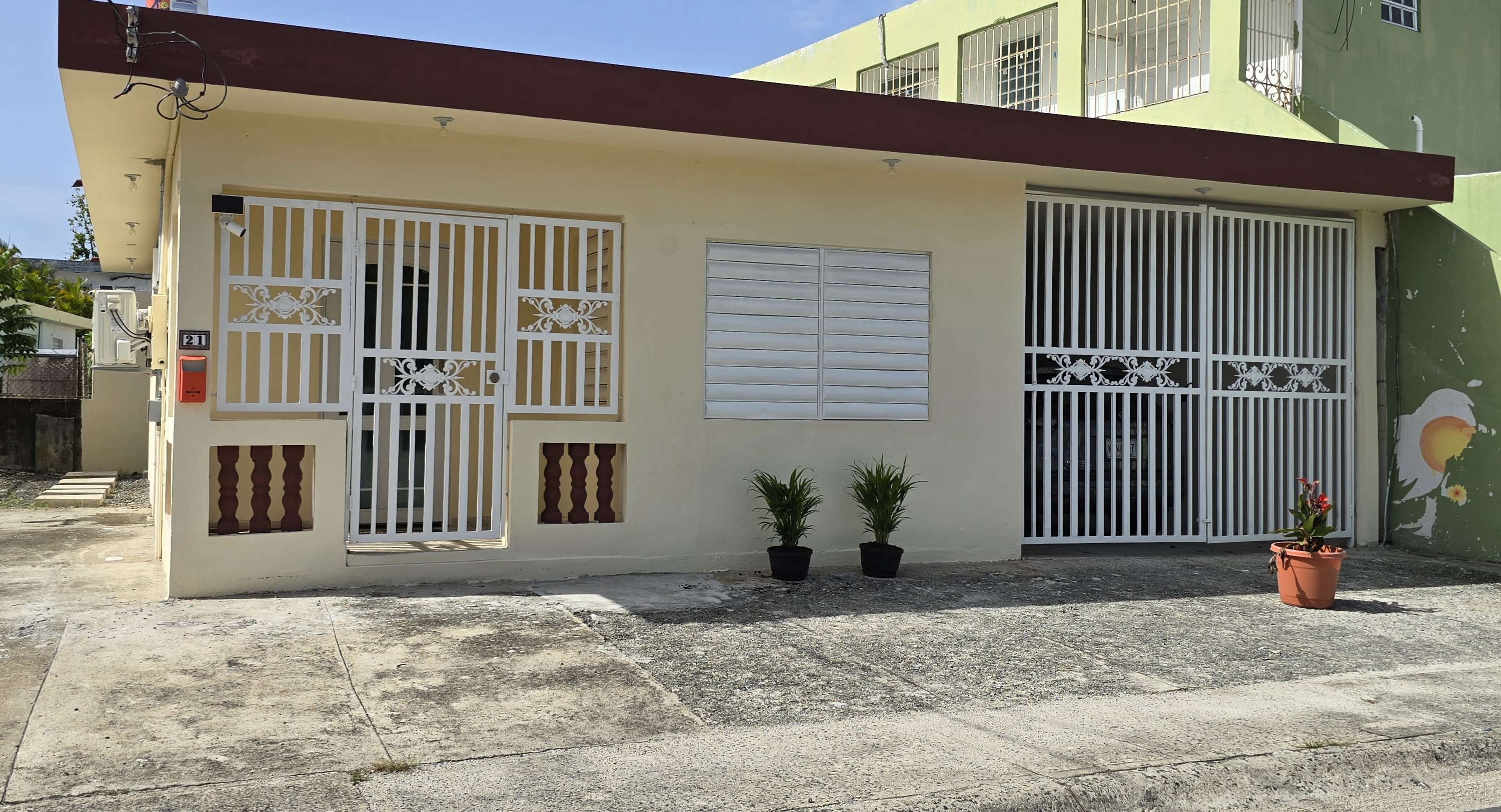 The image shows a modest home with a wrought iron gate and two potted plants in front, situated on a concrete pathway.